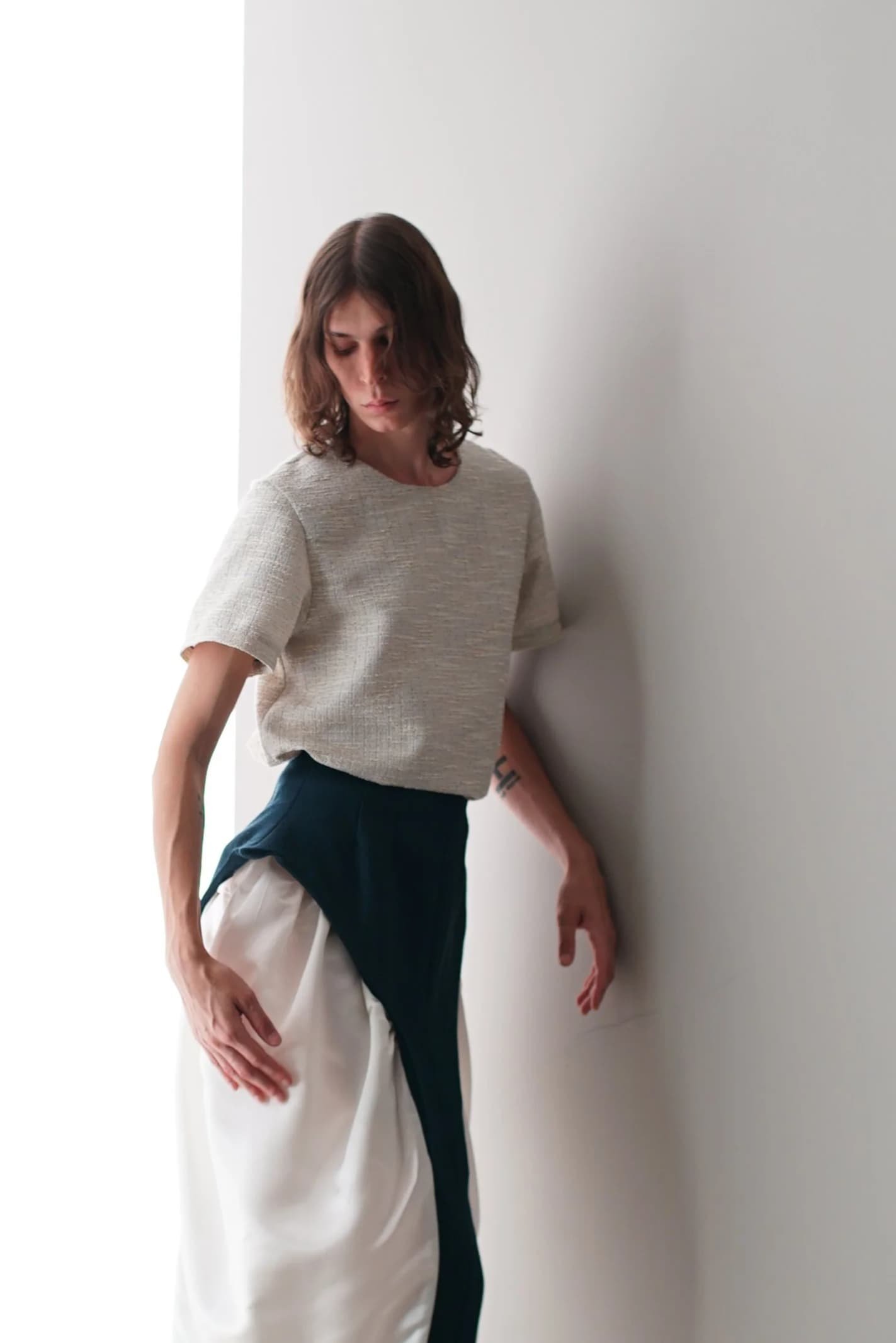 A woman with shoulder-length brown hair standing near a white wall, wearing a light-colored top and a long, layered skirt with white, black, and dark blue sections.
