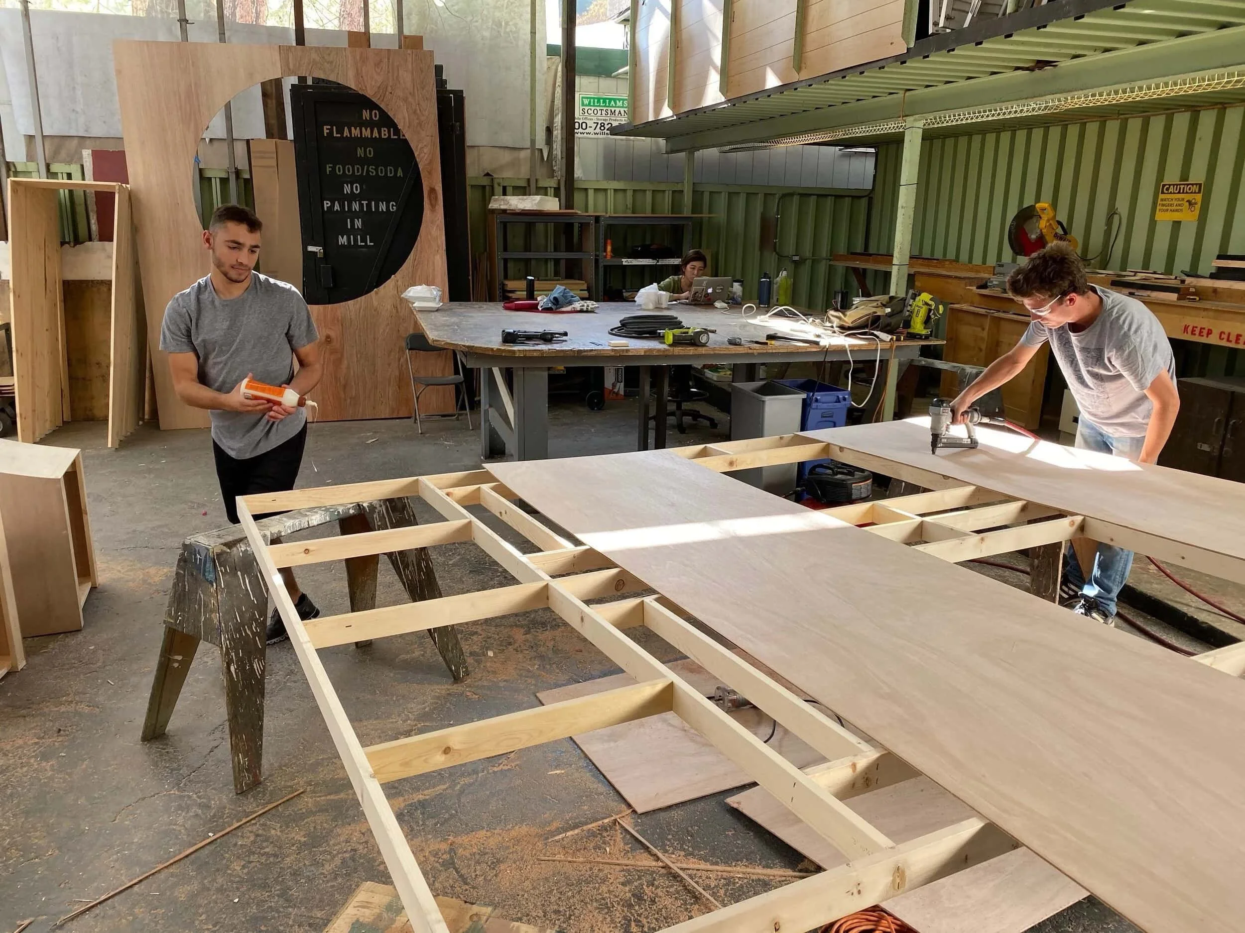 Two men working on woodworking projects in a workshop, one holding a caulking gun and the other using a power drill, with workbenches and tools around.