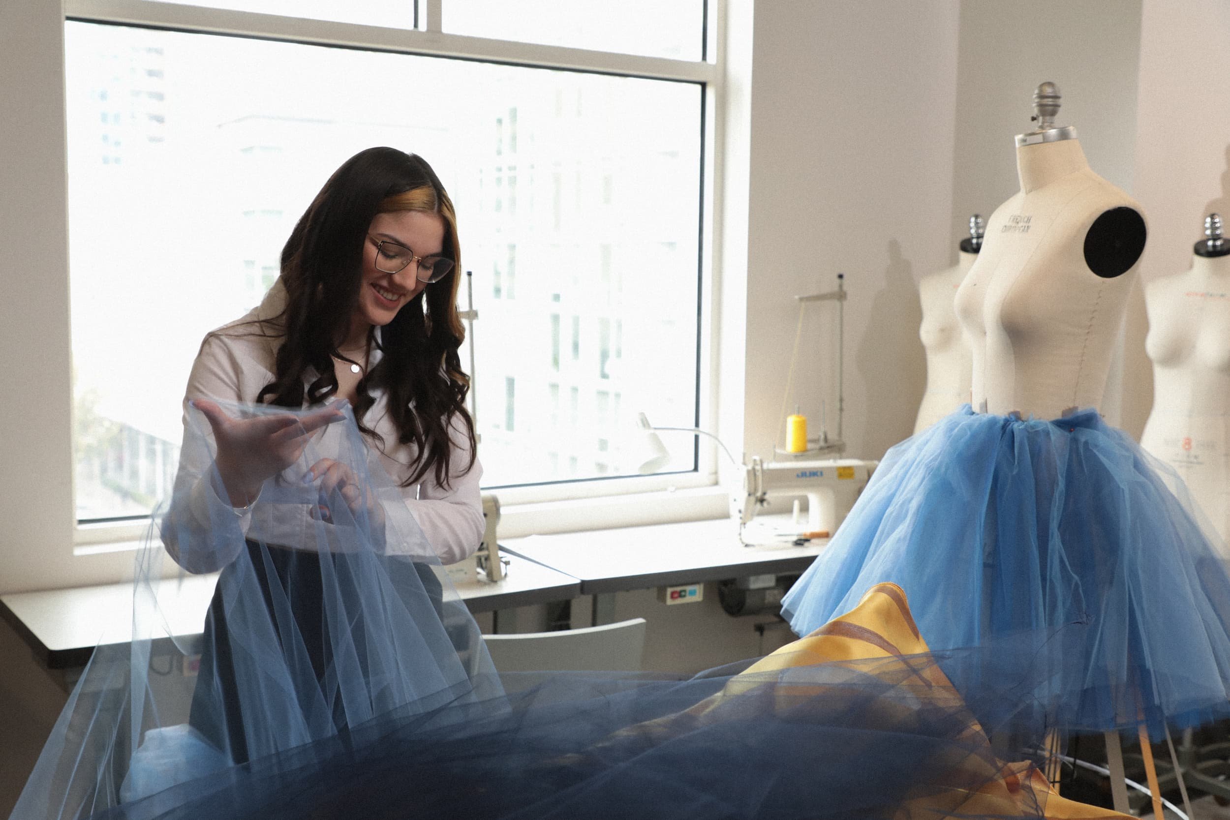 Olivia Bevacqua in a white shirt smiling as she works on a blue tulle skirt inside a sewing studio, with mannequins dressed in similar blue skirts nearby.