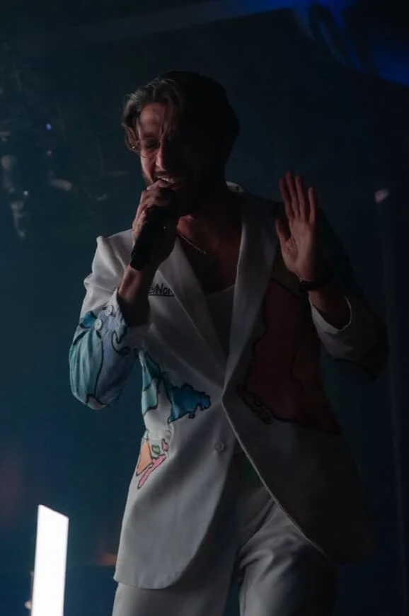 A man singing into a microphone on stage, wearing a colorful jacket, with a dark background and blue lighting.