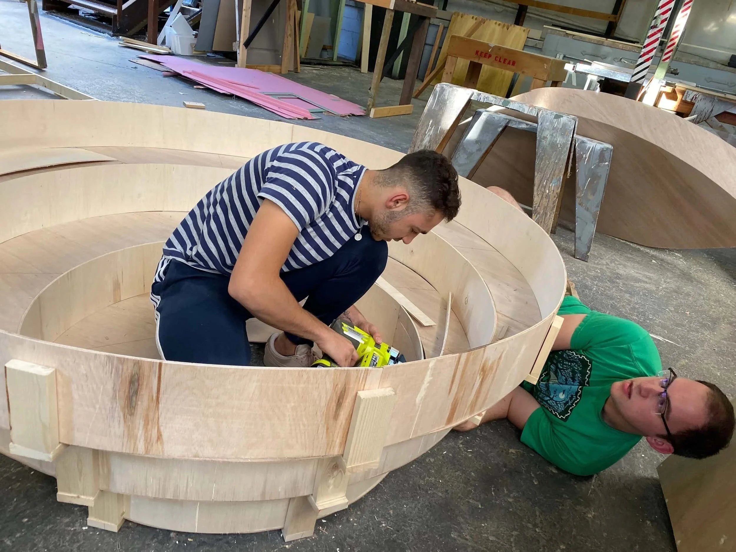 Keagan Roberson and crew working on a wooden circular structure inside a workshop, one kneeling and using a power tool, the other lying on the floor beside it.