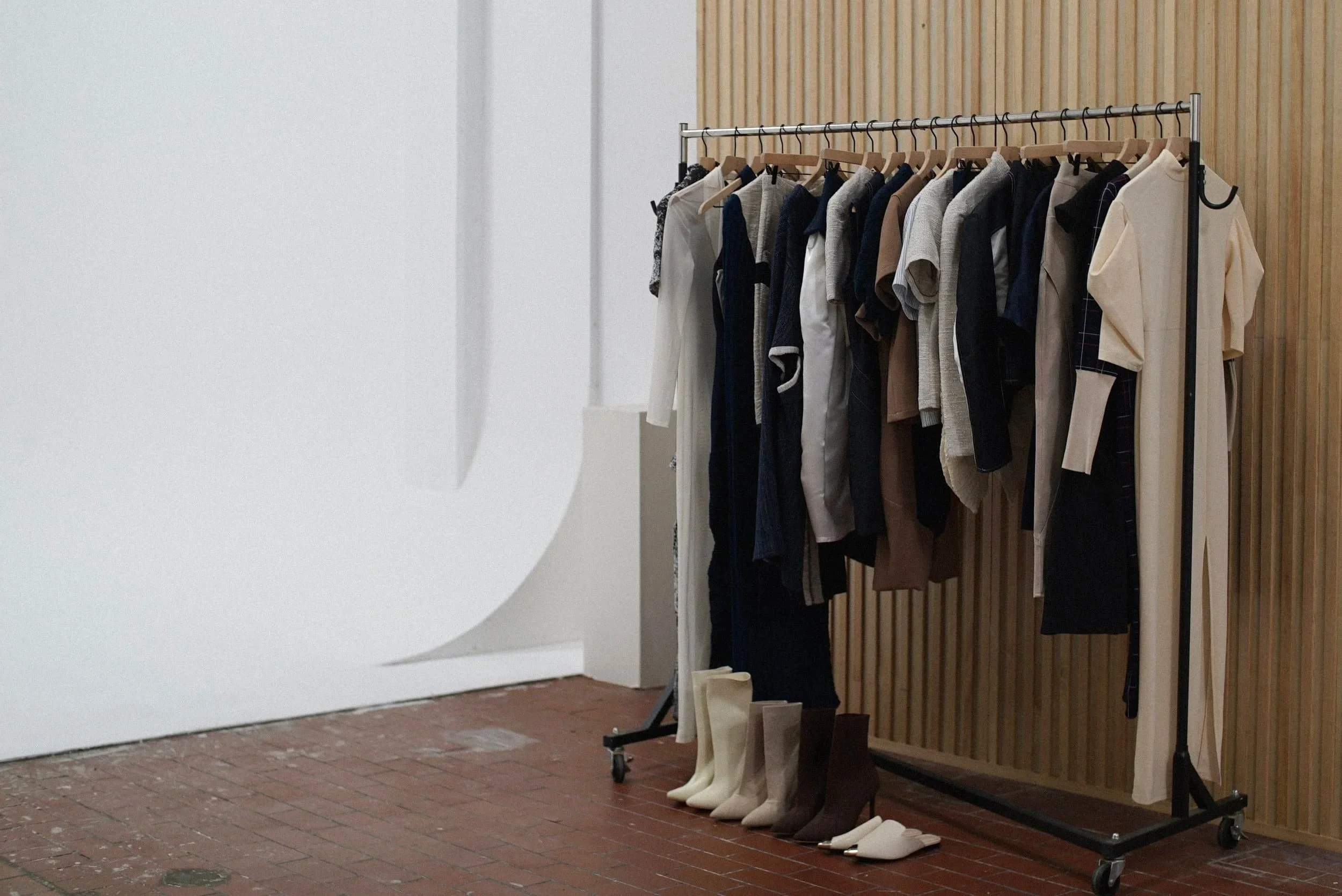Clothing rack with various shirts, pants, and dresses, and pairs of boots and slippers on the floor in a room with a wooden wall and brick floor.