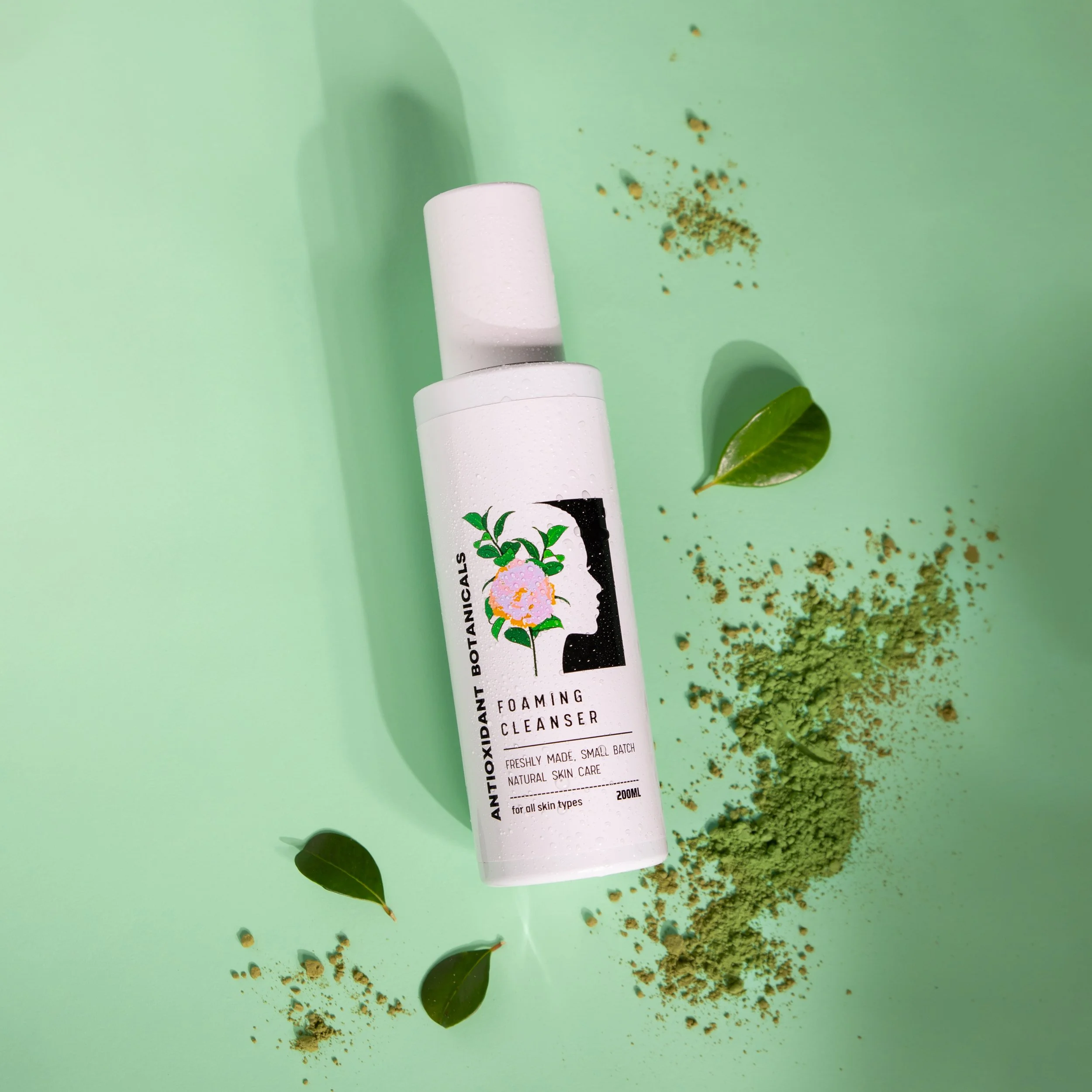 A white bottle of foaming cleanser with botanical illustrations on a light green background, surrounded by green powder, leaves, and small scattered particles.