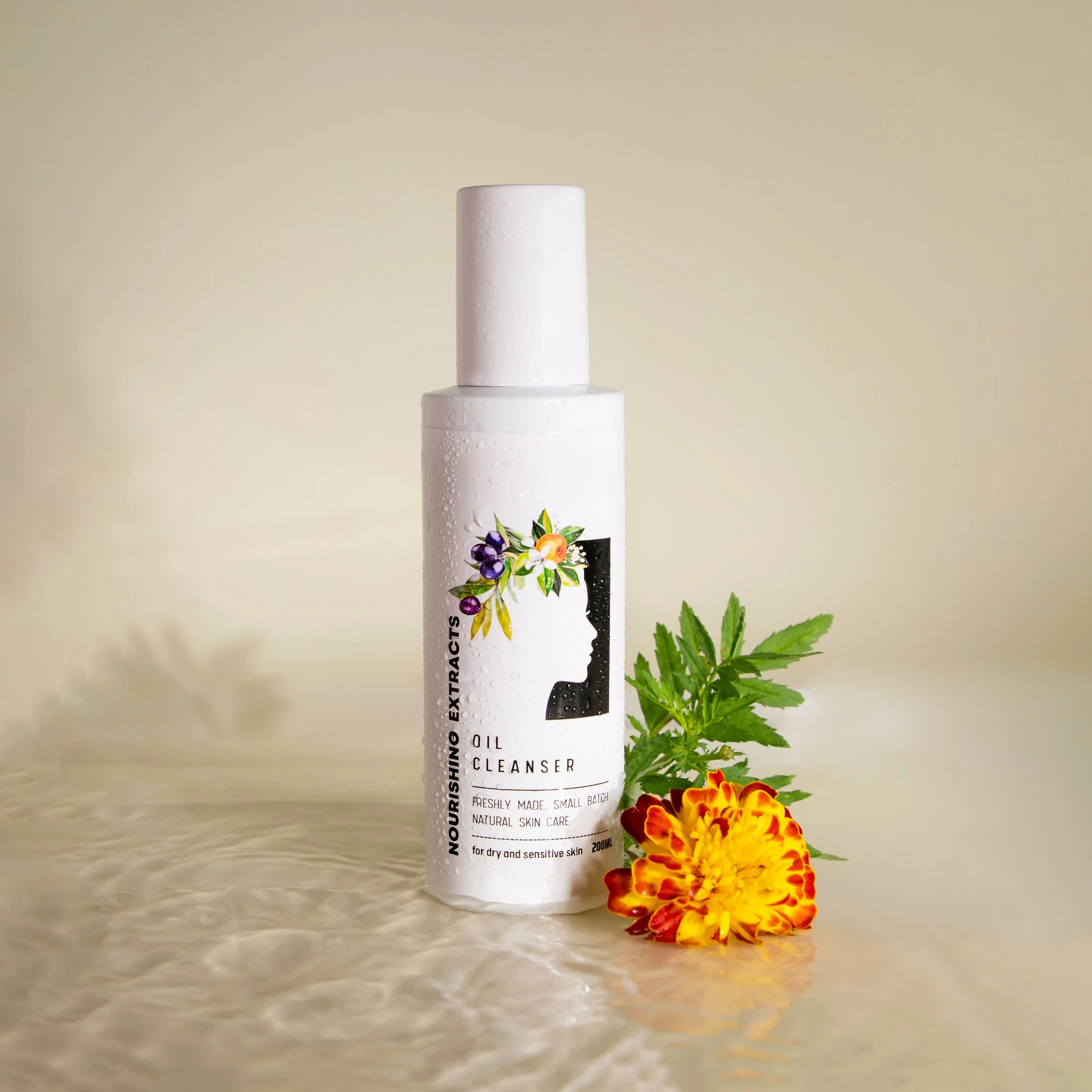 A white bottle of Nourishing Extracts Oil Cleanser with a floral label, placed on a reflective surface next to green leaves and a yellow and orange marigold flower on a neutral background.