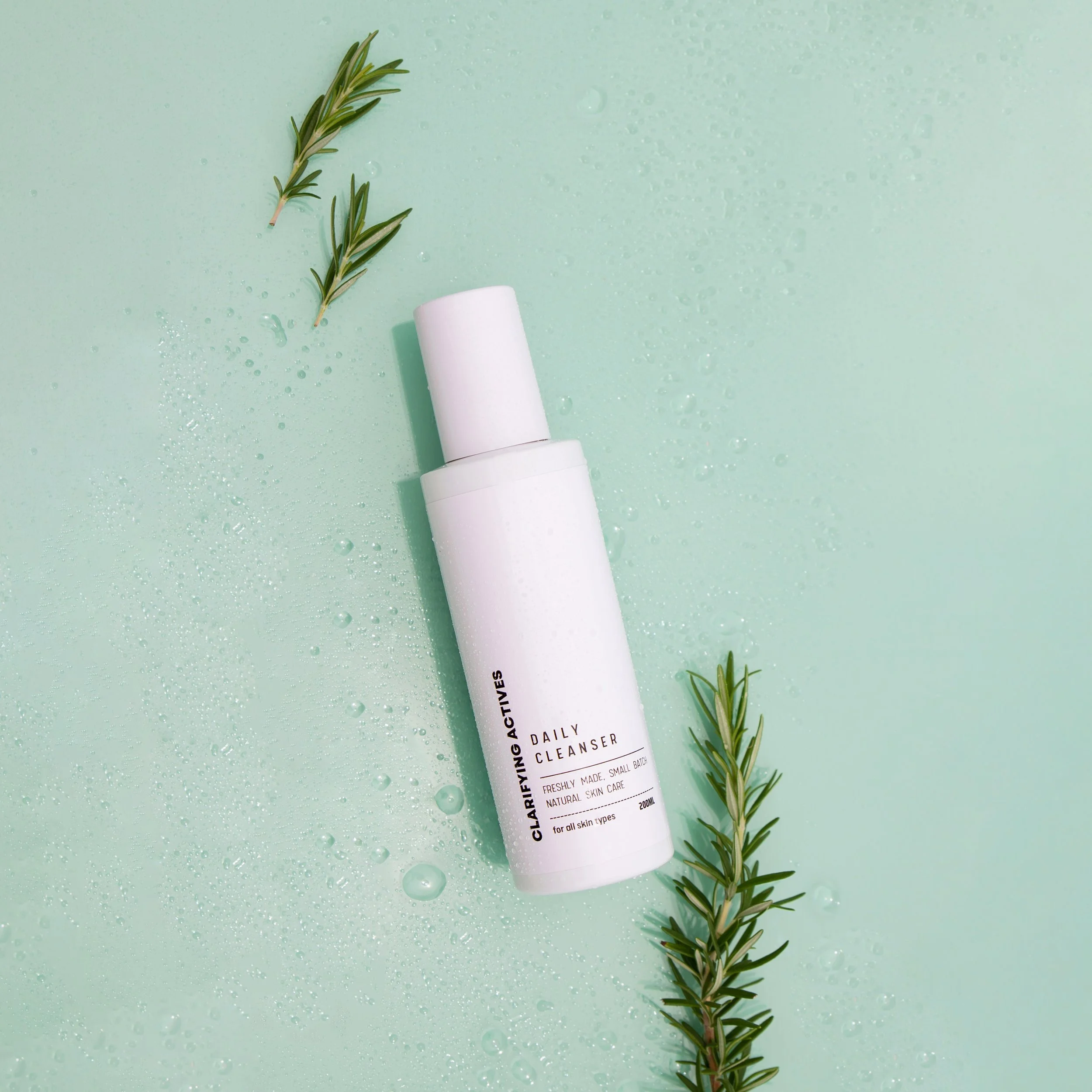 White bottle of daily cleanser lying on a pale green surface with water droplets and two sprigs of rosemary.