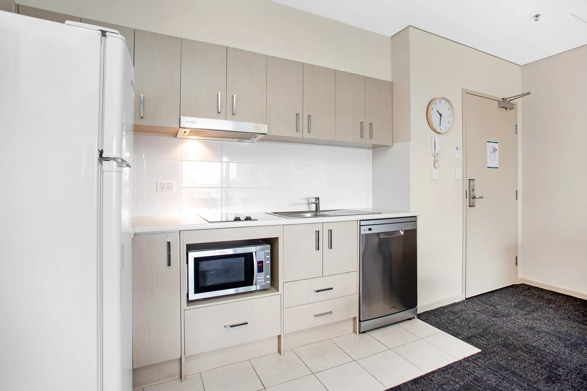 Modern kitchen with light wood cabinets, white countertops, built-in microwave, electric cooktop, stainless steel dishwasher, and a white refrigerator. Wall clock and intercom present.