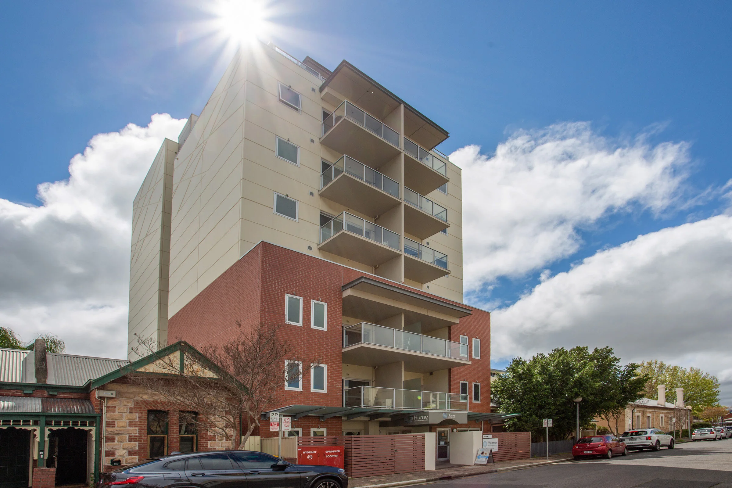 Modern multi-story apartment building with balconies under a bright sun, surrounded by parked cars on an urban street.