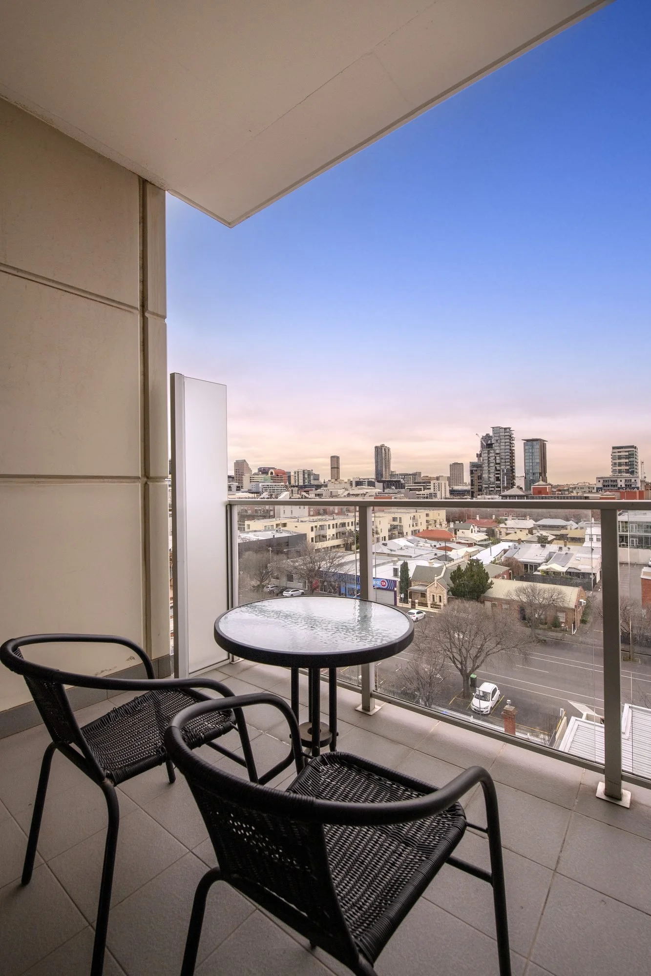 Balcony view overlooking a cityscape with trees, rooftops, and hills in the background, featuring a small round glass table and two black chairs.