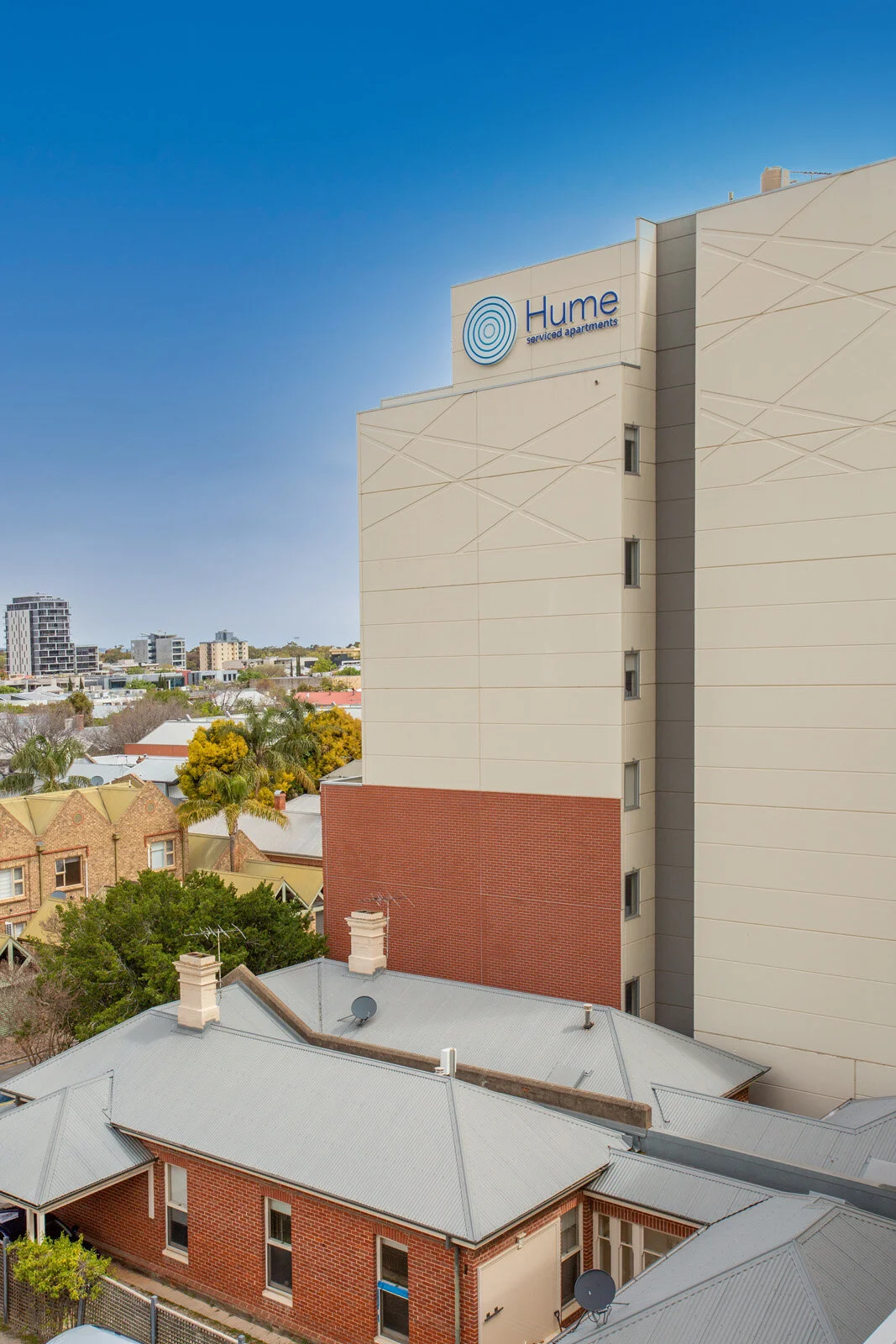 View of a tall building labeled "Hume serviced apartments" with a blue logo, surrounded by residential buildings with metal roofs and brick exteriors, against a clear blue sky.