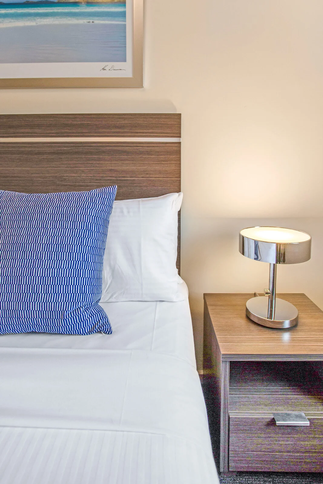 Modern bedroom with a wooden headboard, blue and white striped pillow, crisp white sheets, bedside table with a sleek lamp, and framed beach artwork.