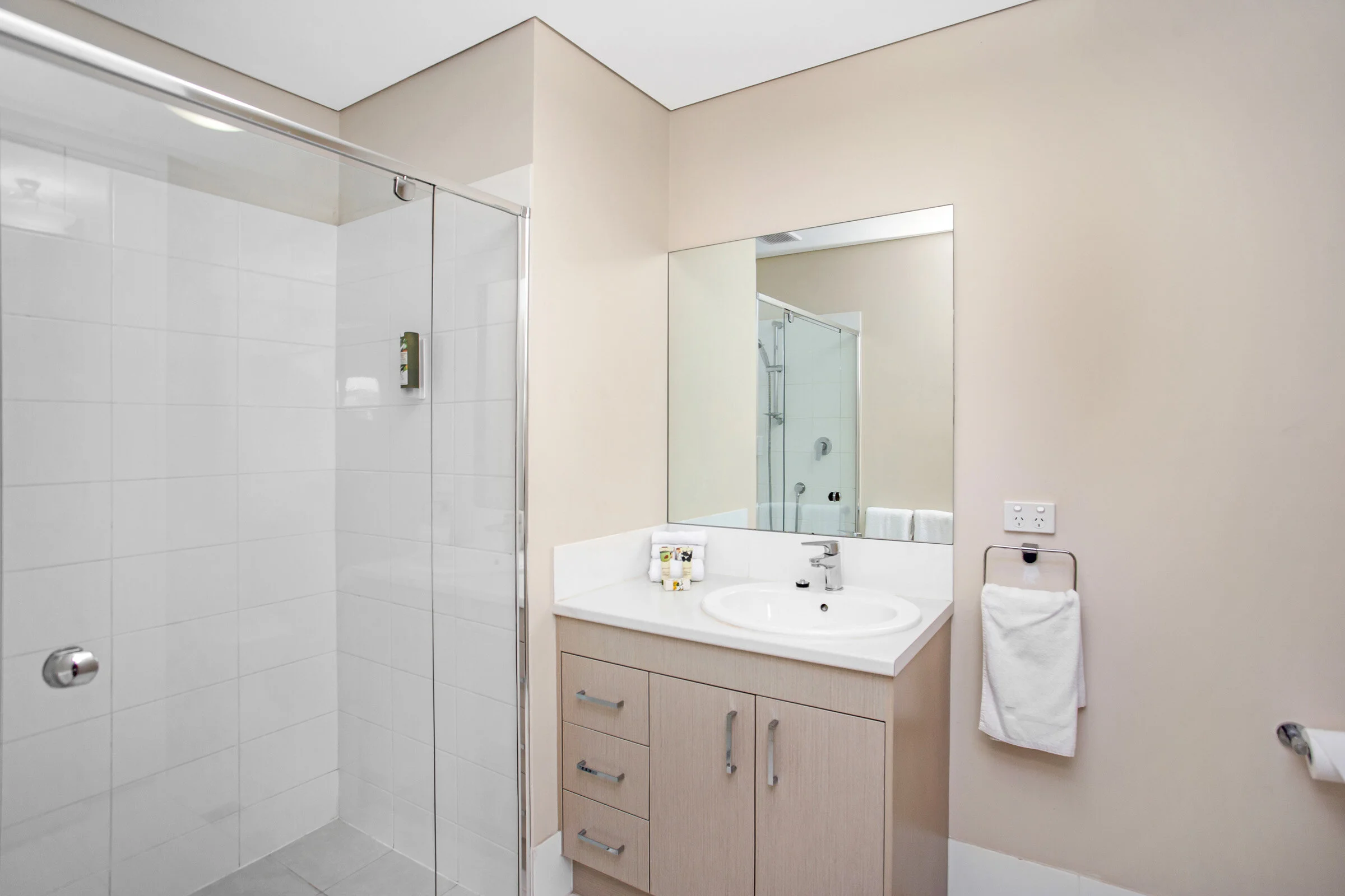Modern bathroom with glass-enclosed shower, beige walls, large mirror, wooden vanity with sink, and towel rack.