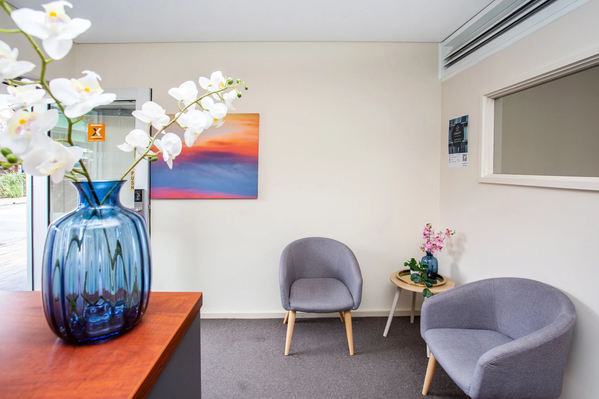 Modern waiting room with gray chairs, a wooden table with pink orchids, wall art, a large window, and a blue vase with white flowers.