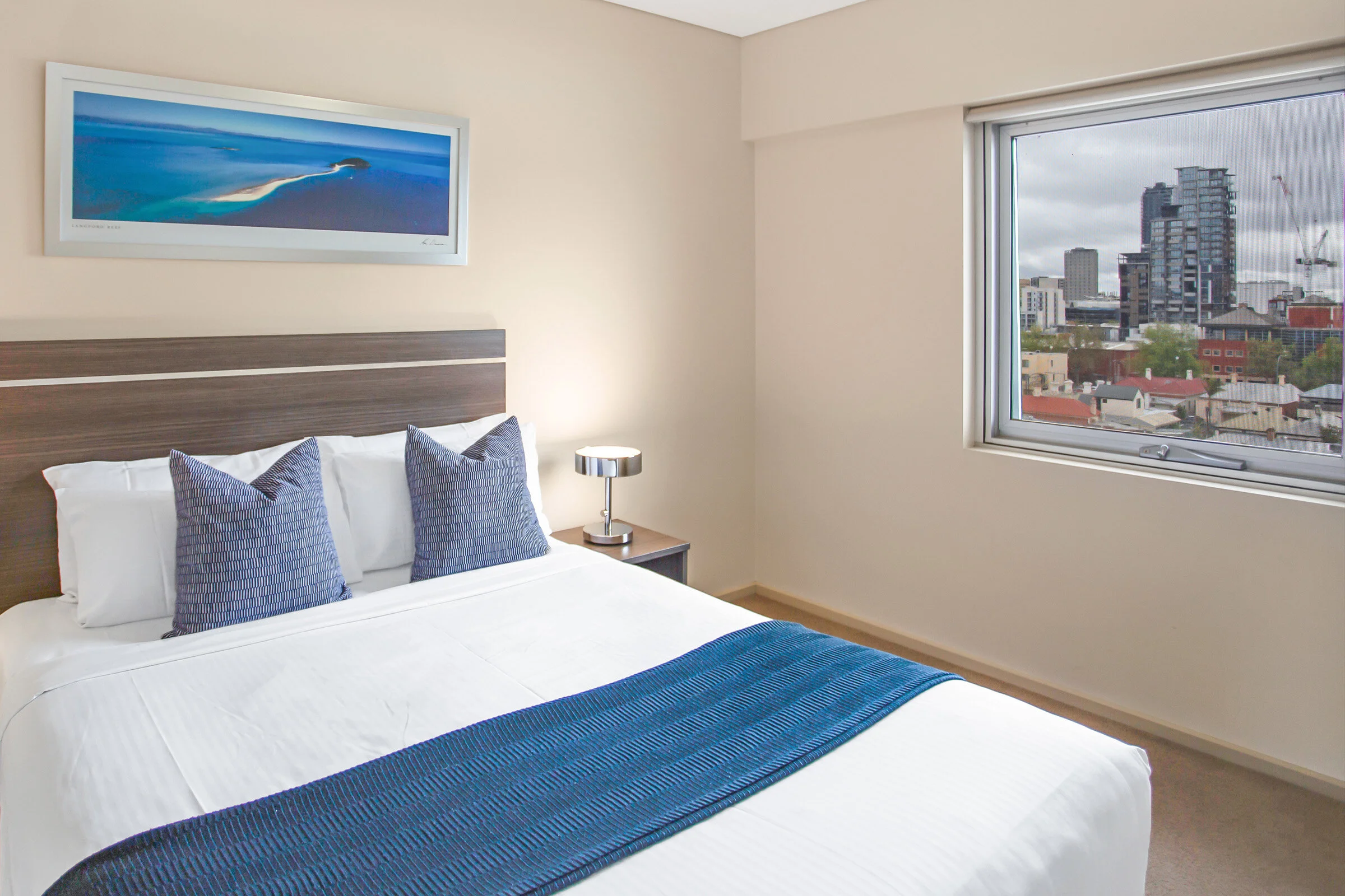 Modern hotel room with a large bed, blue pillows and throw, nightstand with a lamp, framed seascape photo on the wall, and window with city view.