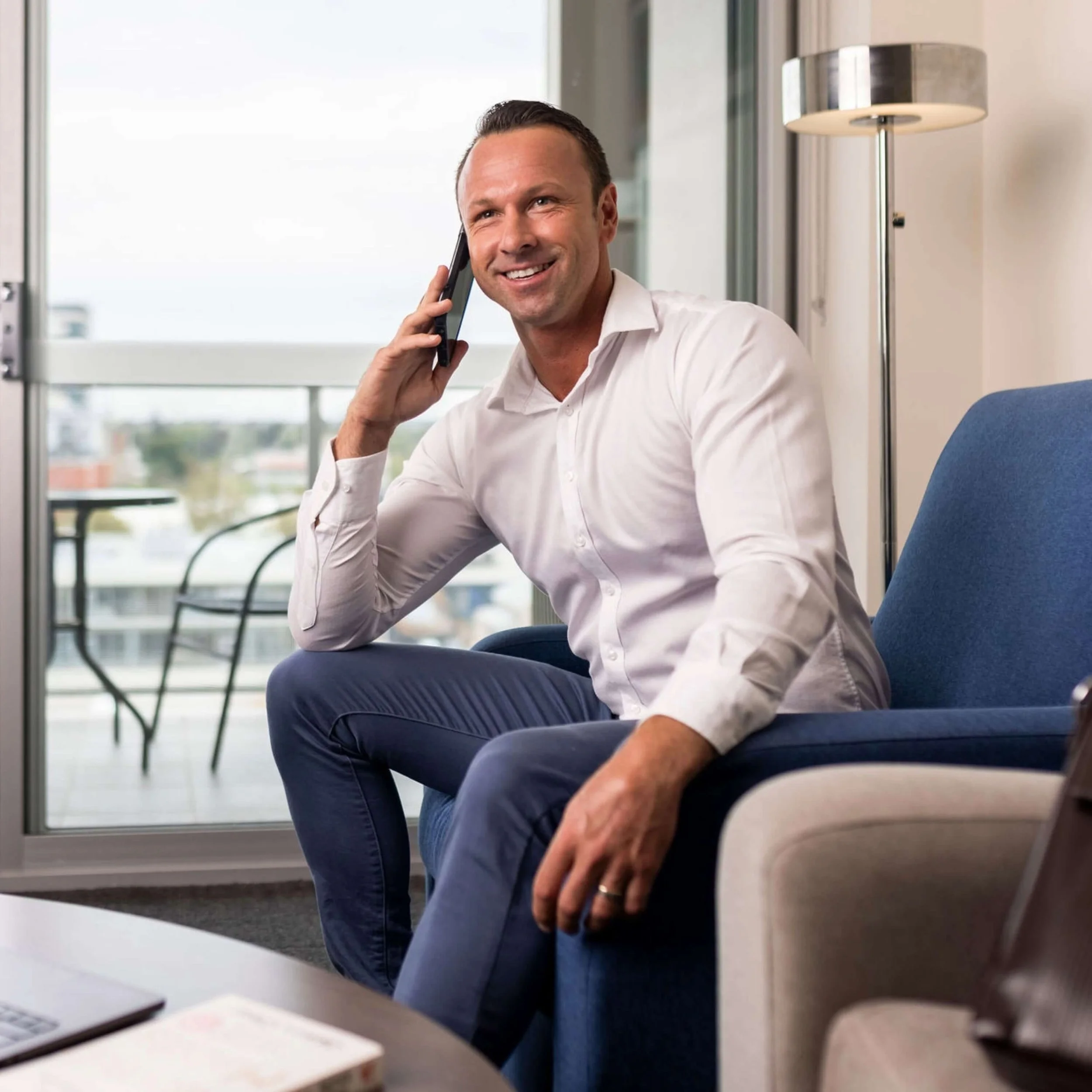 A man in a white shirt and blue pants sitting on a blue couch, smiling while talking on a mobile phone, in a well-lit room with a sliding glass door leading to a balcony.