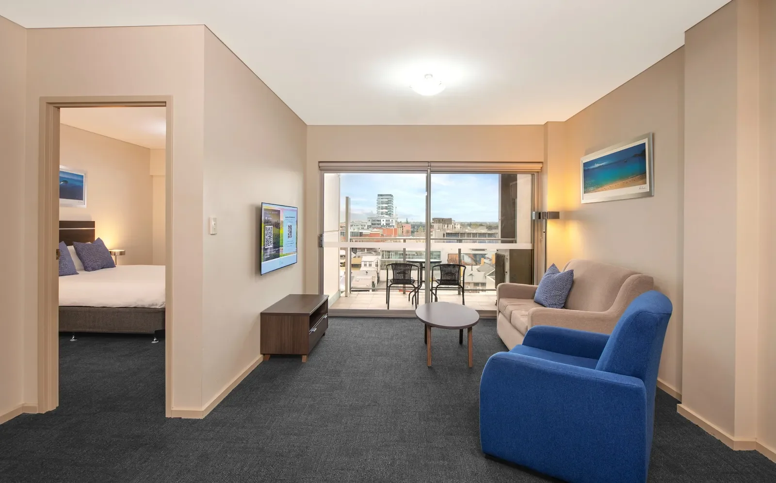 Living room with beige sofa, blue armchair, small round coffee table, and wall-mounted TV, with a balcony and city view in the background.