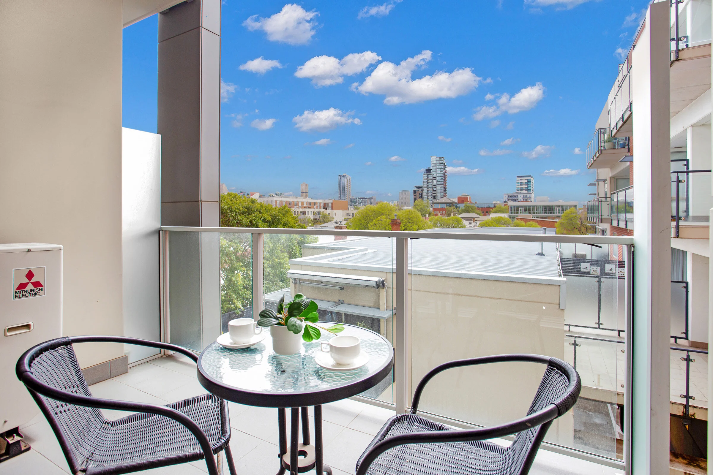 Balcony with two chairs and a table, city view, blue sky, white clouds, plant on table, and outdoor AC unit.
