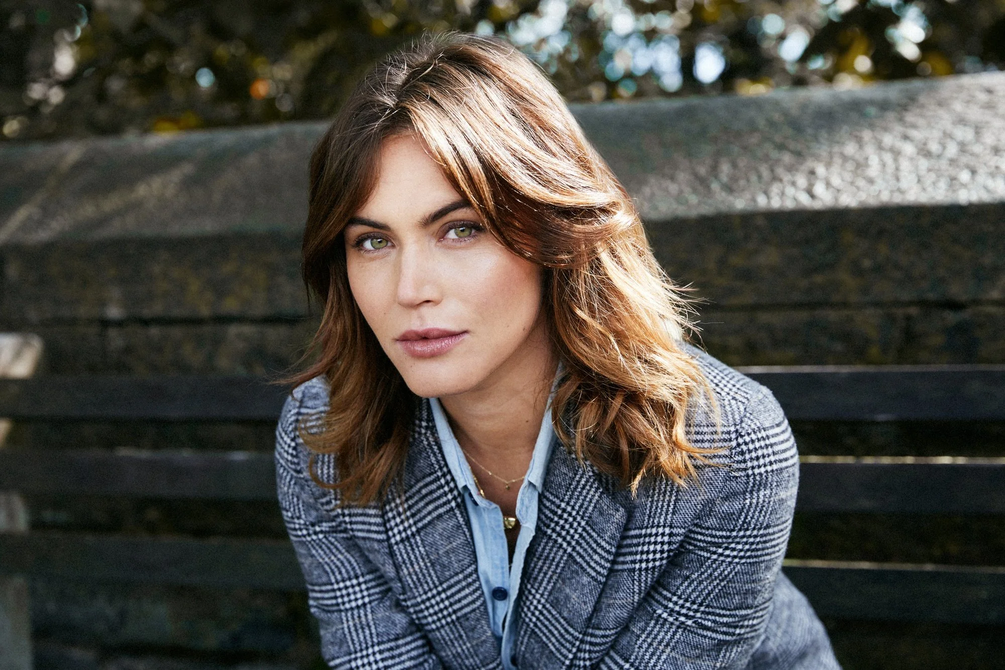 Portrait of a young woman with brown hair sitting on a park bench in the fall. She is backlit with soft, even natural light covering her face. She has green eyes and is wearing a grey, black &  white plaid coat with a denim button down shirt.