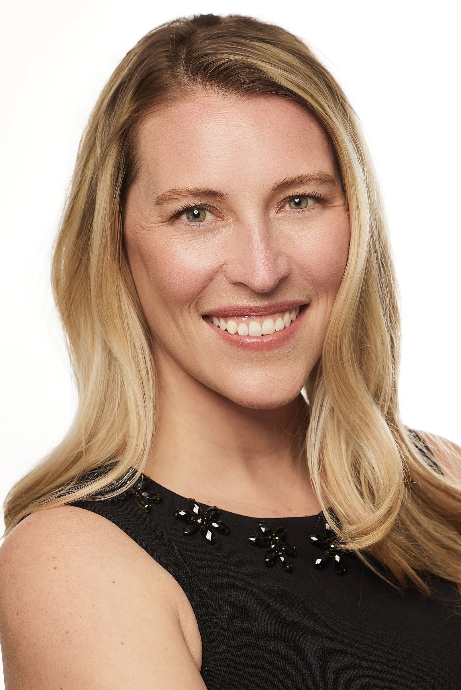 Close-up of a woman with blonde hair, smiling, wearing a black sleeveless top with black floral embellishments on the collar, against a white background.
