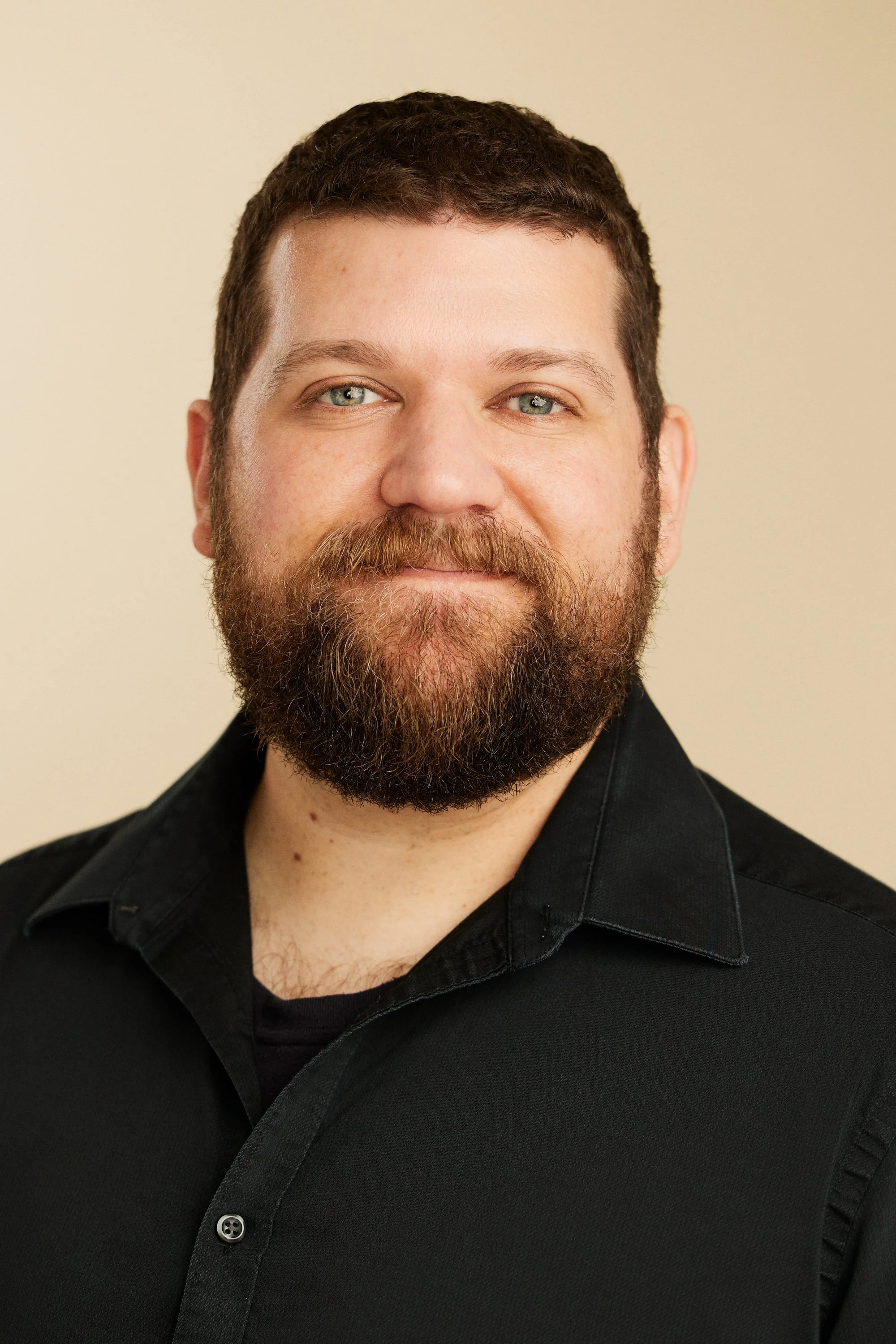 A man with light skin, red hair, and a beard, wearing a turquoise blazer, smiling with his head resting on his hand, against a light turquoise background.