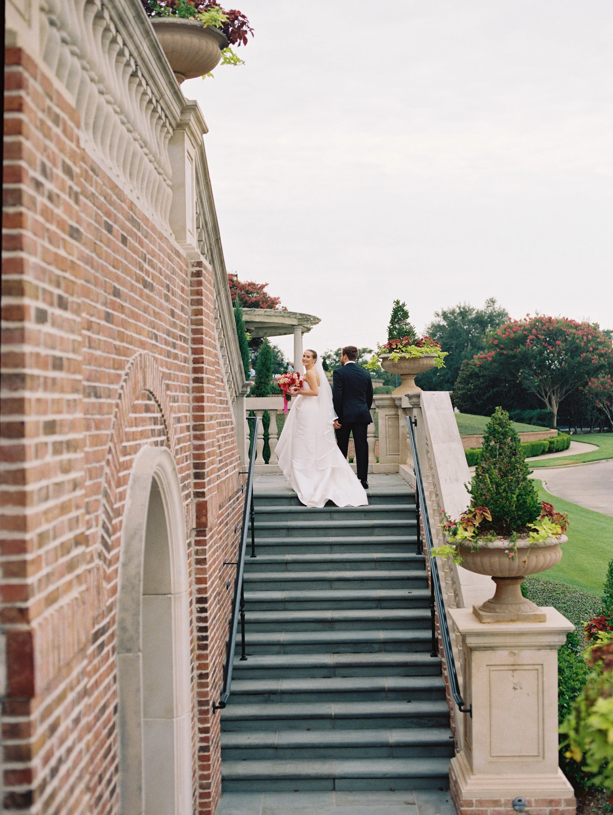 Outdoor Texas wedding photo outside St. Maryβs Catholic Church in College Station, Texas, with groom kissing bride on the cheek as her veil blows in the wind and the wedding party cheers, captured by a College Station wedding photographer.