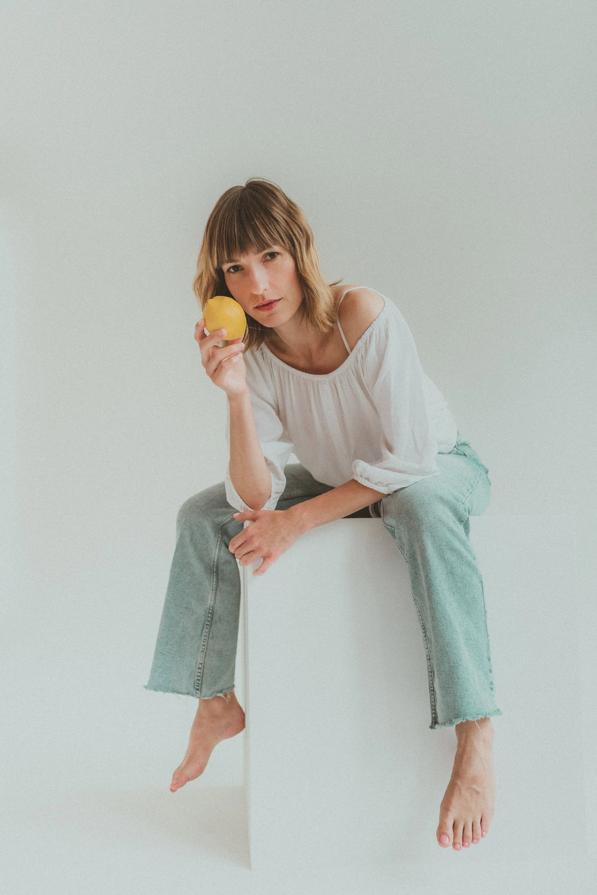 A woman with shoulder-length brown hair and bangs, wearing a white off-shoulder top and light blue jeans, sitting on a white block against a plain light-colored background, holding a yellow apple close to her face.
