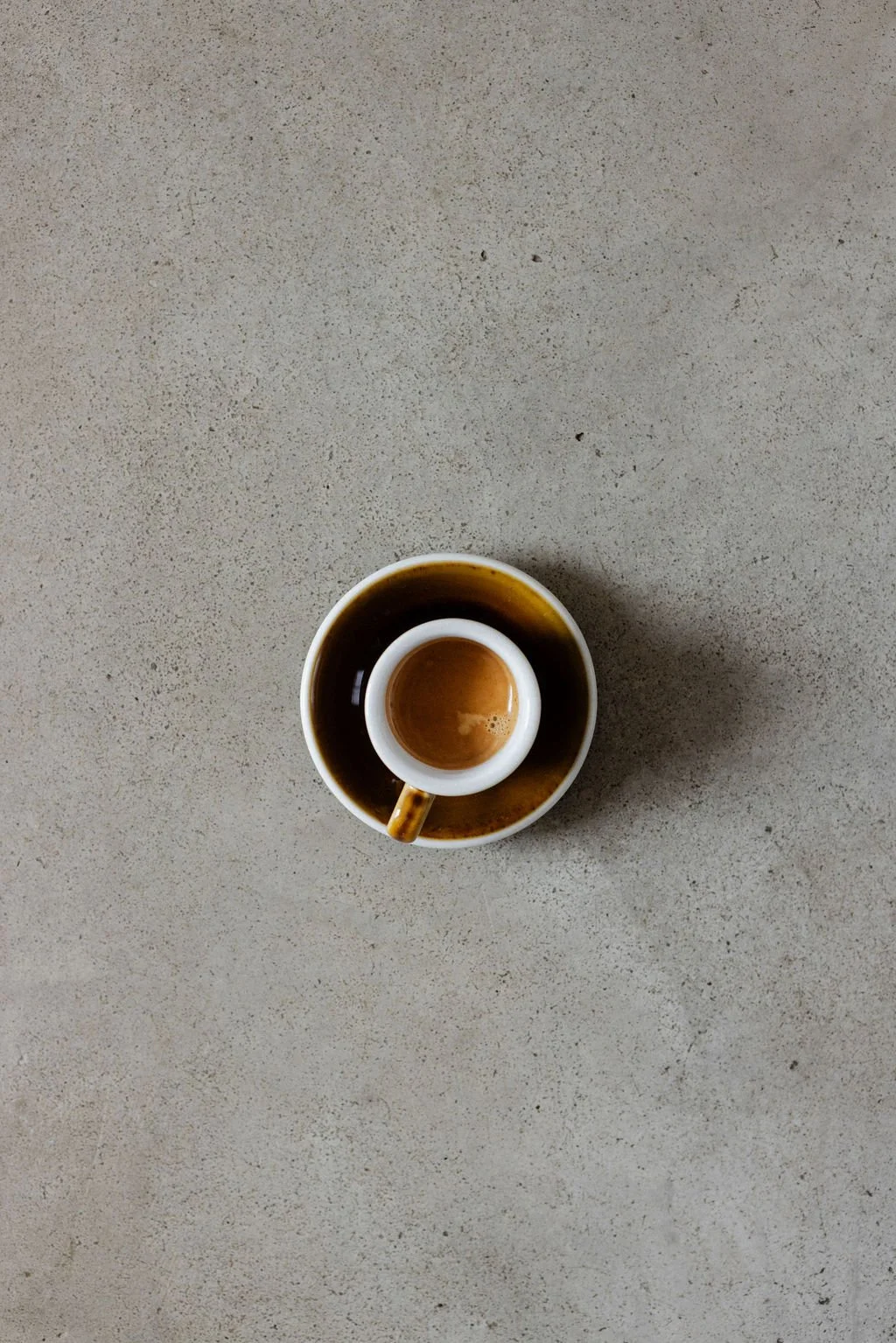 A small cup of espresso sitting inside a saucer, viewed from above on a light-colored, textured surface.