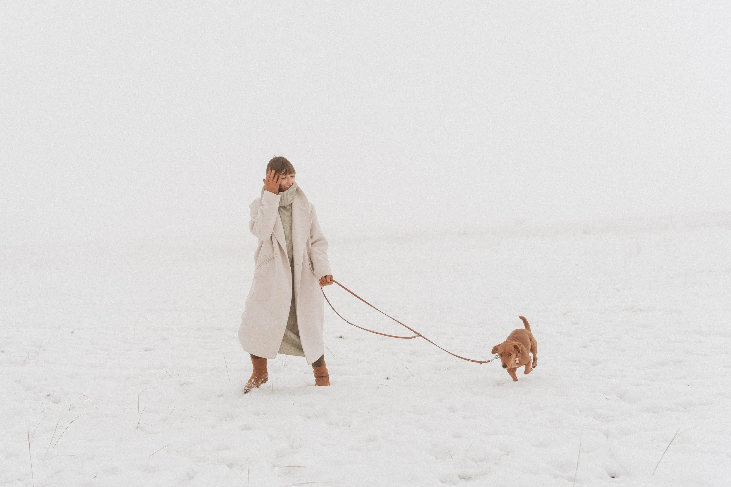 A woman with light brown hair wearing a beige coat, turtleneck, and boots walking a dog on a leash in a snowy landscape.