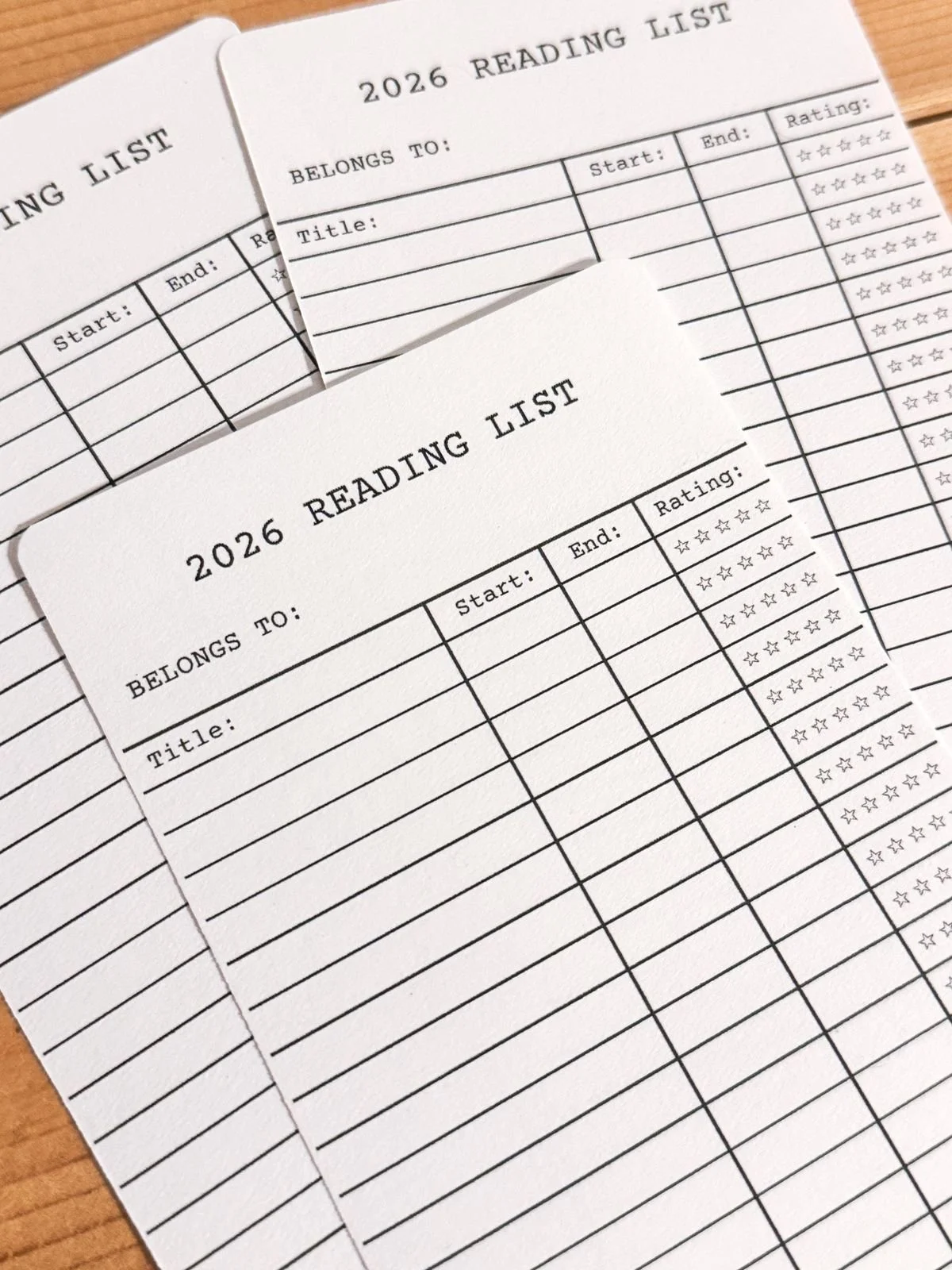 Two printed reading list sheets for 2026, with columns for book titles, start and end dates, and ratings, resting on a wooden surface.