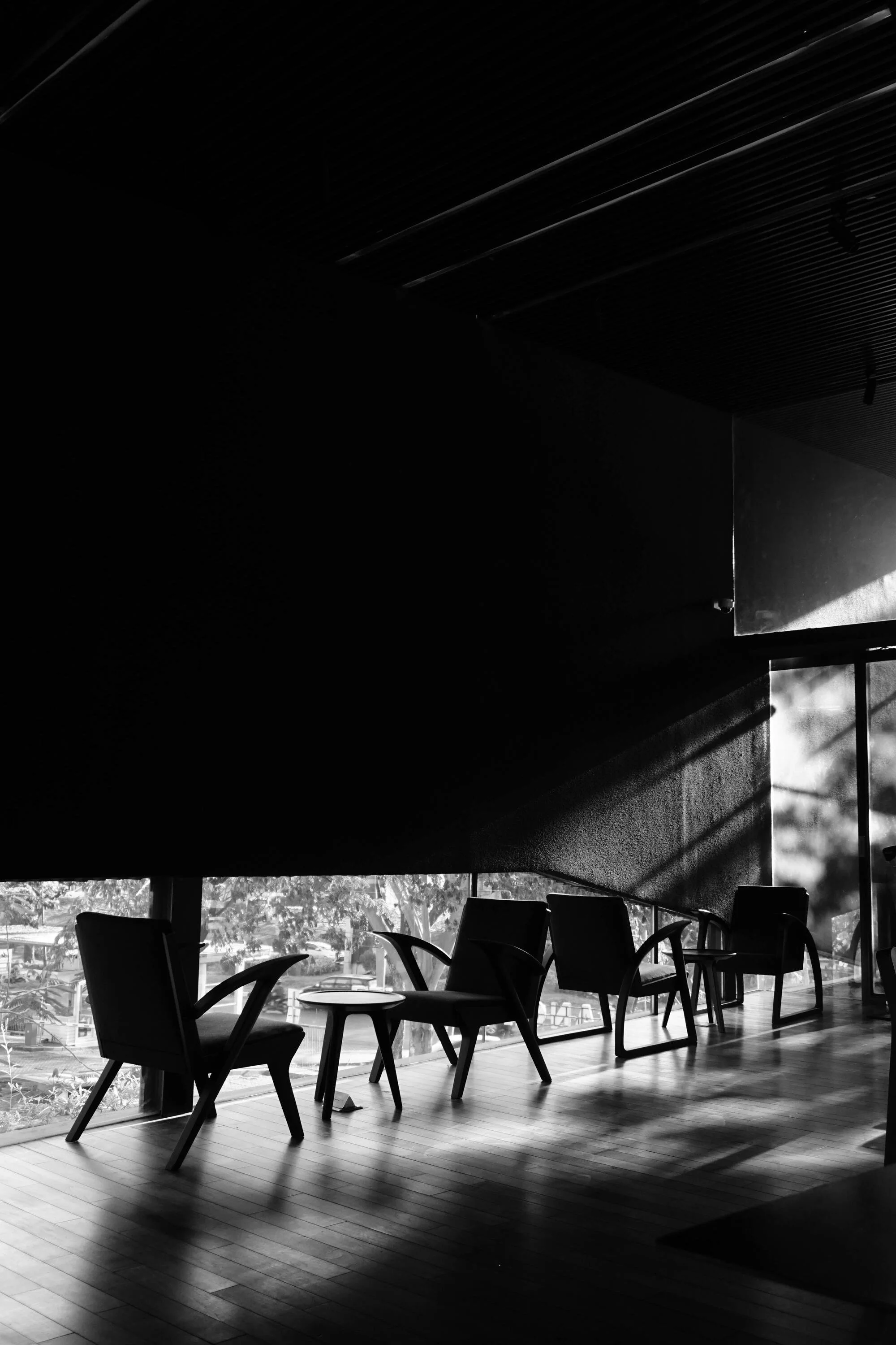 Interior of a modern cafe or restaurant with wooden floors, chairs, and tables positioned near large windows. Sunlight streams through the windows, creating shadows inside the space, and the ceiling is made of a dark, slatted material.