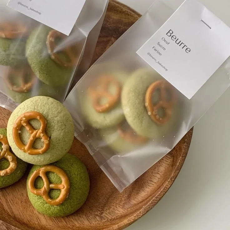 Green cookies with pretzel-shaped caramel decorations on a wooden platter, some in clear plastic bags labeled 'Beurre' with French ingredients.