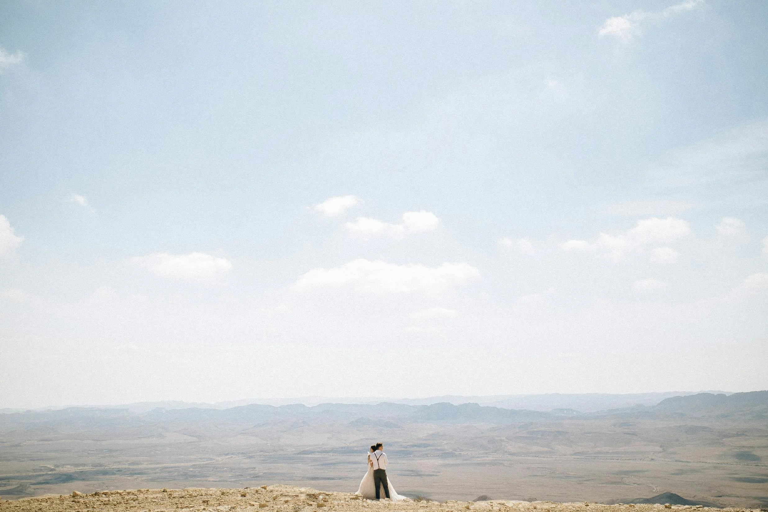 Wedding couple embracing on a desert landscape with distant mountains and a clear blue sky.