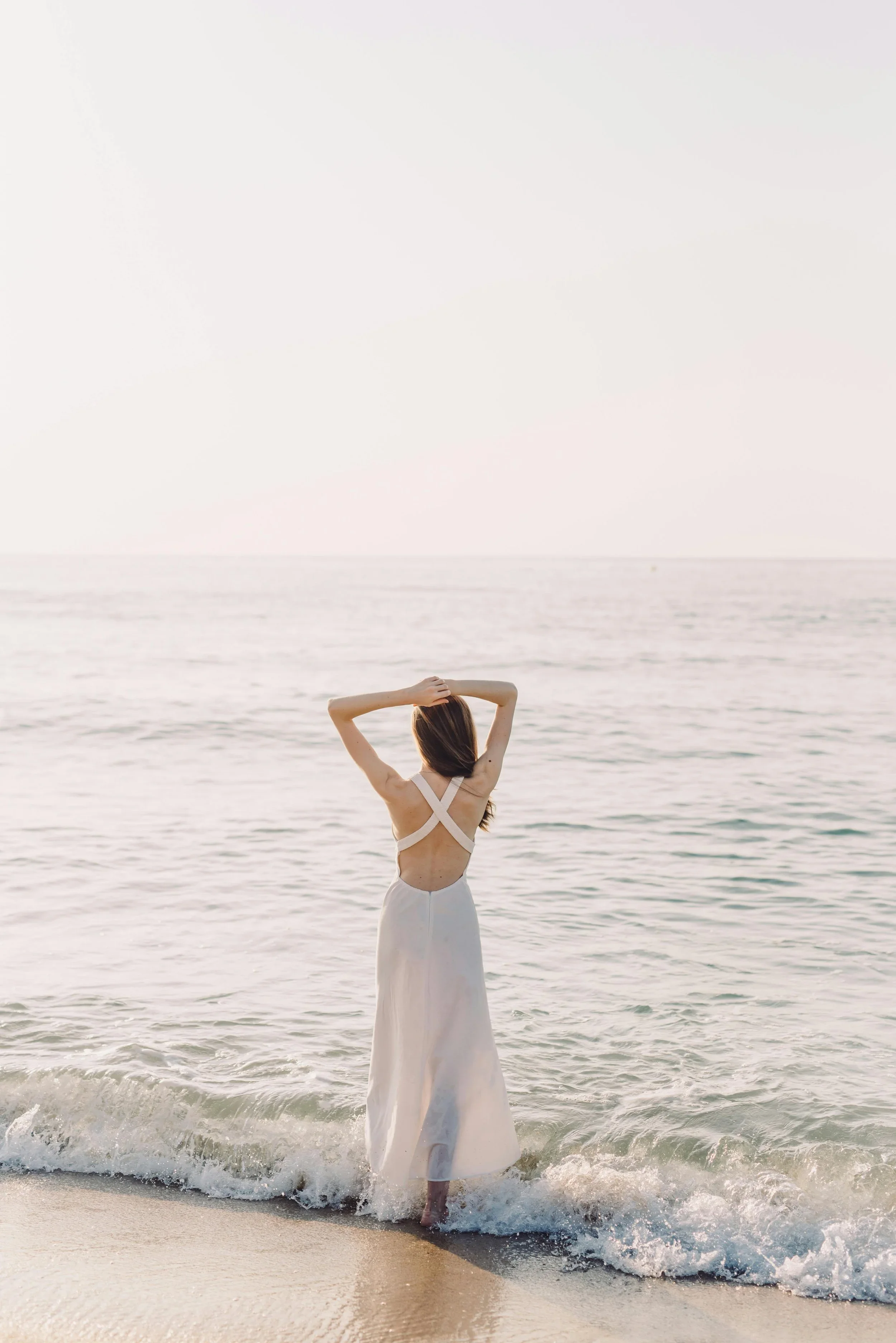 A woman in a white dress standing in the ocean with her back to the camera, her arms raised above her head, at the shoreline with gentle waves and a cloudy sky.