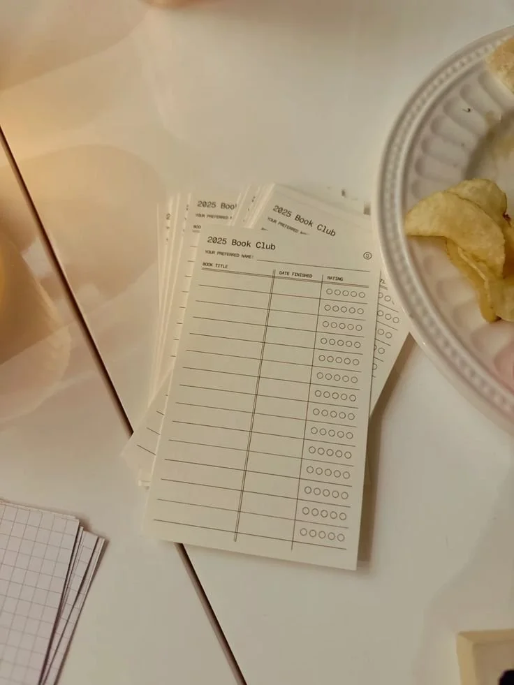 Stack of papers or forms titled '2025 Book Club' on a white table, with a part of a plate of potato chips and a transparent cup in the upper right corner.