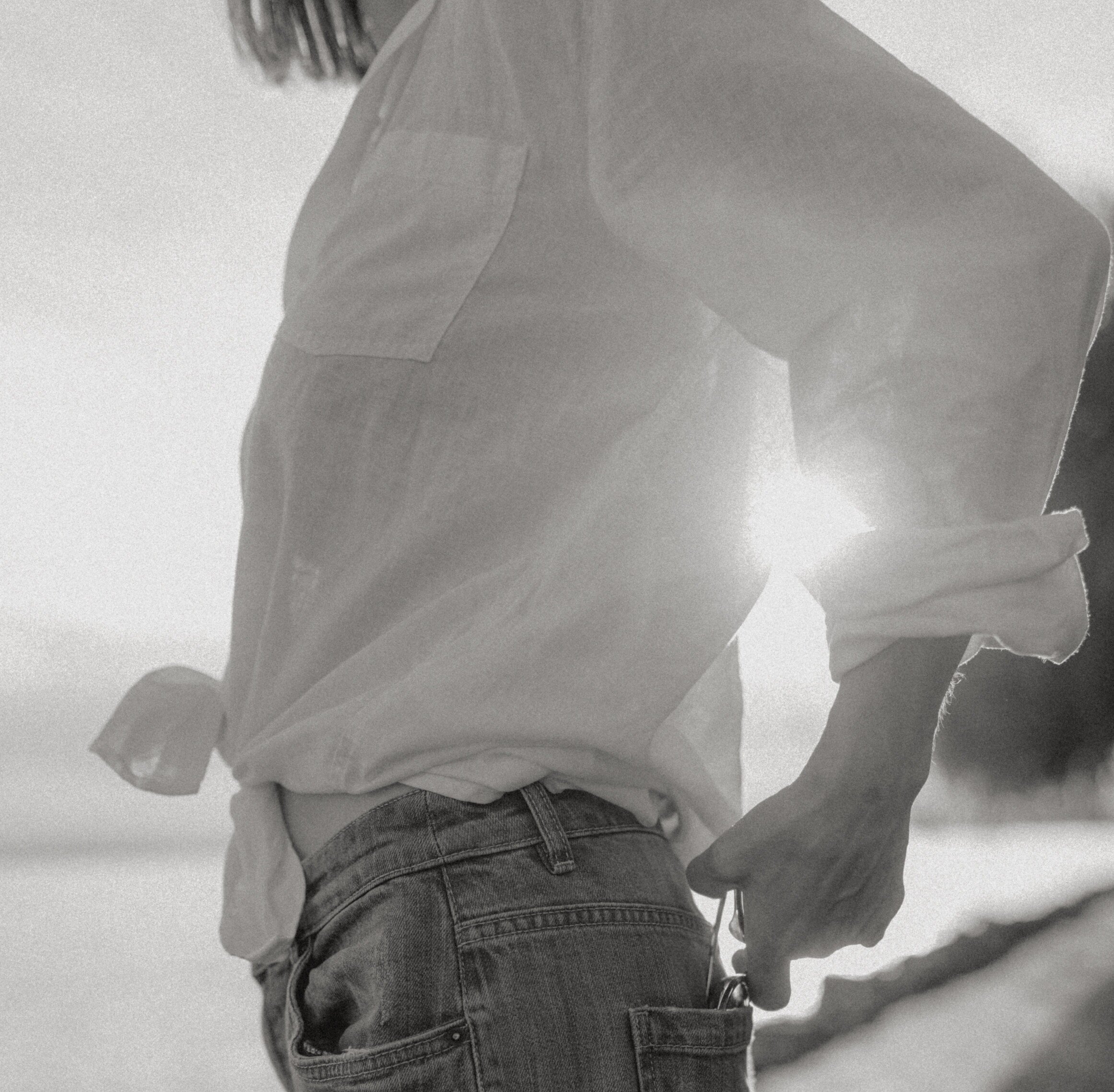 A person wearing a light jacket and jeans standing against a plain wall, with their face partly visible and a casual pose.