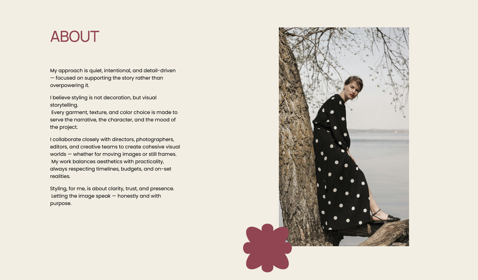 A woman in a black polka dot dress and black shoes sitting on a tree branch by the water with bare tree branches above her, during daytime.