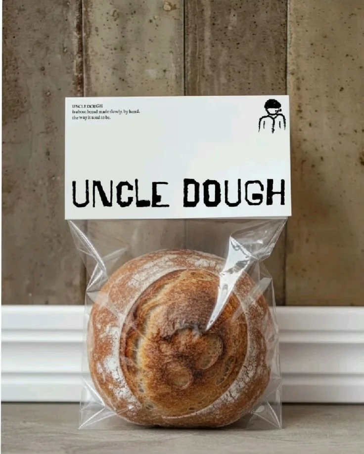 A wrapped cinnamon roll in a clear plastic bag with a white paper label reading 'Uncle Dough' and a small illustration of a person wearing glasses and a backpack, set against a wooden background.