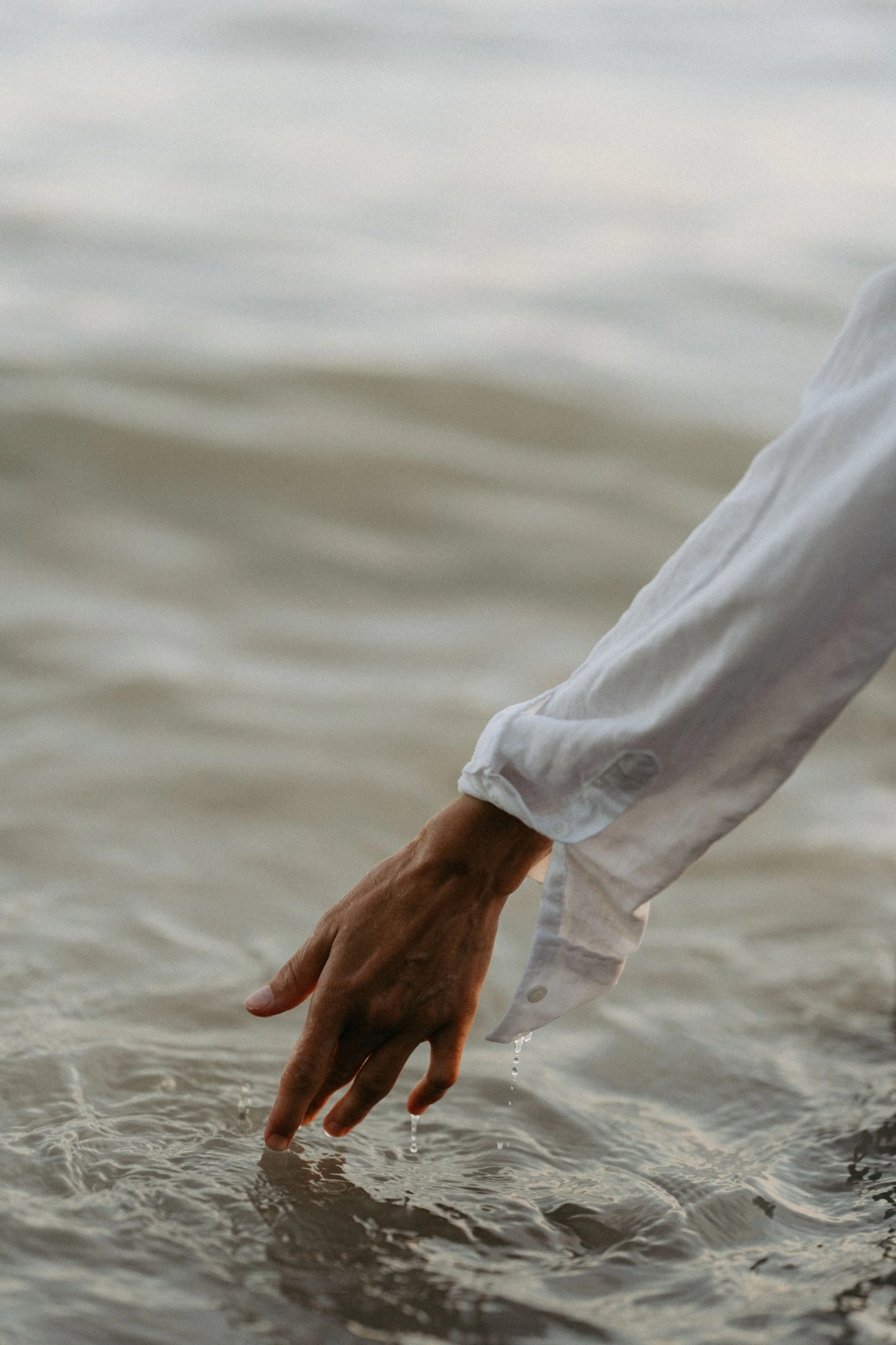 Close-up of a person's hand and arm reaching into the water at the beach, wearing a white long-sleeve shirt, with water dripping from their fingers.