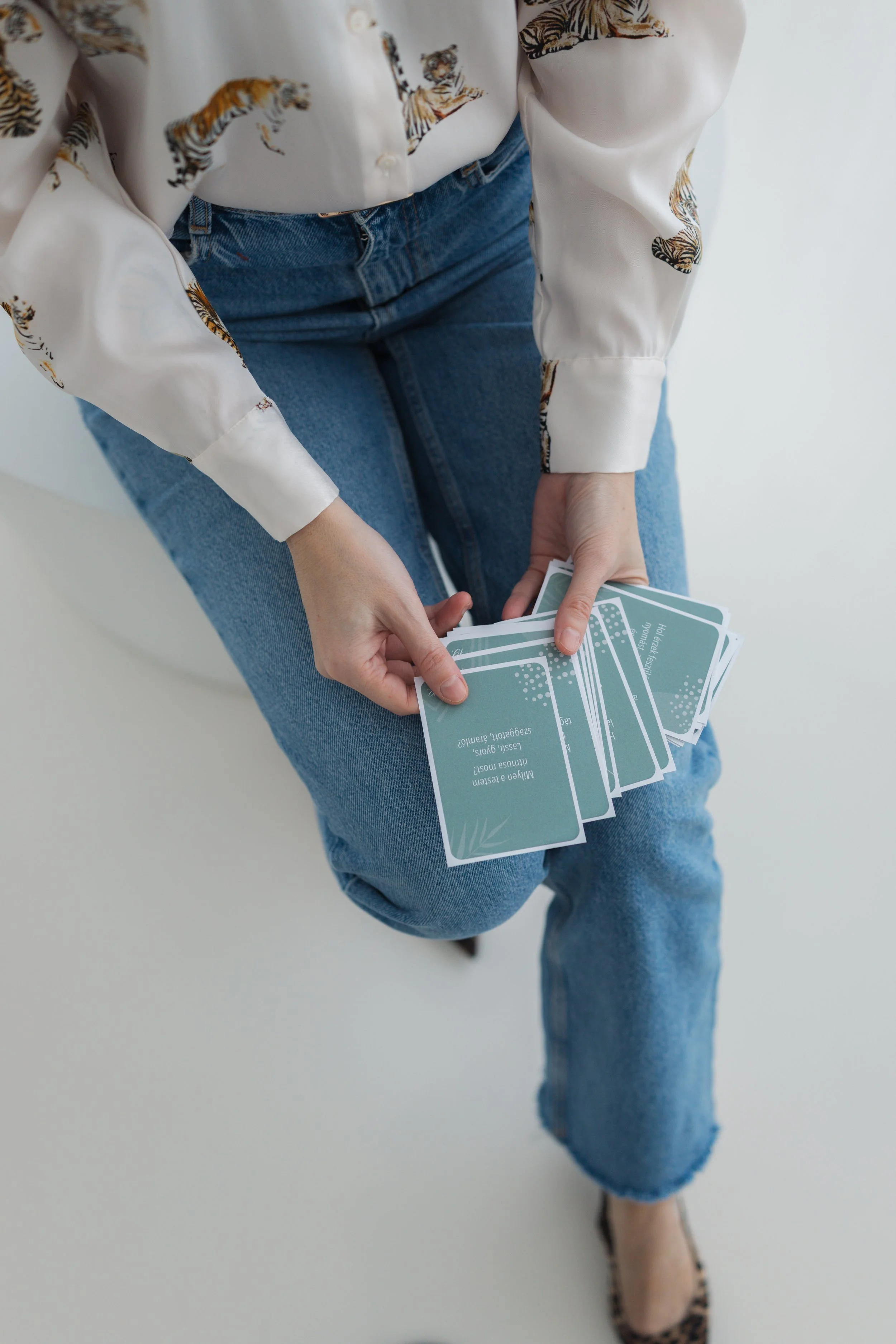 Person wearing a white blouse with tiger print and blue jeans, holding a handful of light blue cards with white text, and wearing leopard print shoes.