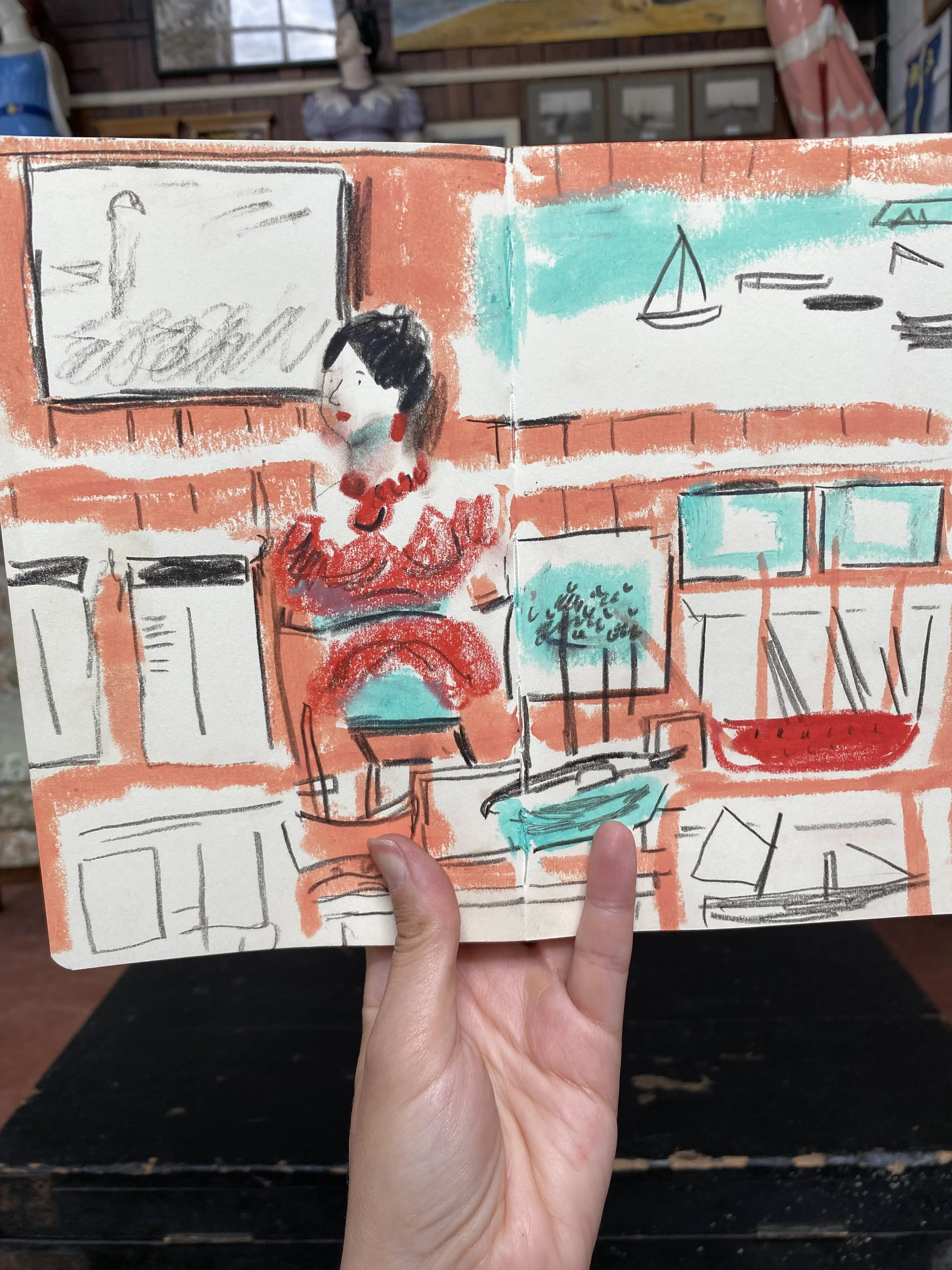 A hand holding a sketchbook with a colourful drawing of a figurehead from a boat, surrounded by framed pictures on a wall