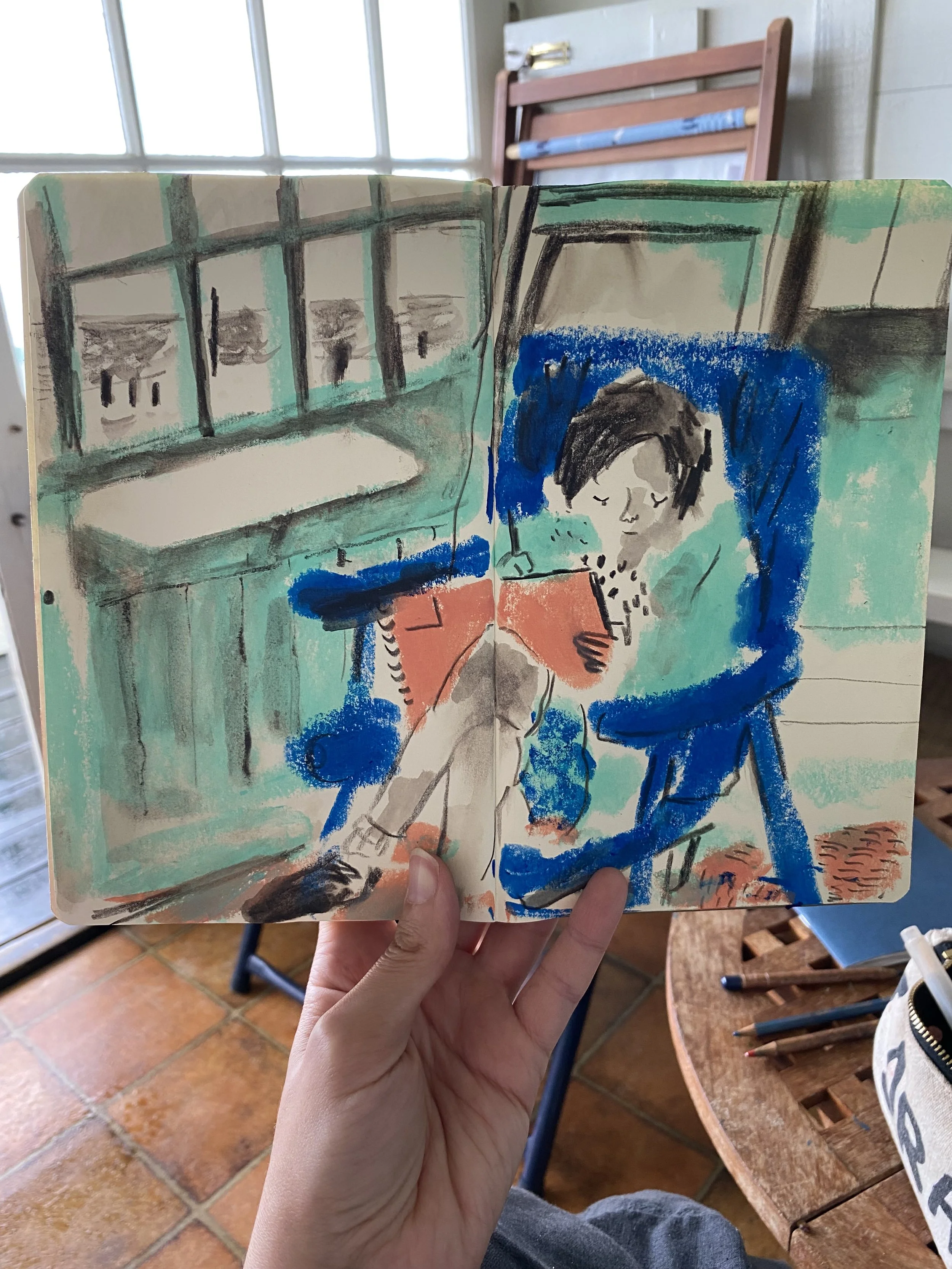 A hand holding a sketchbook with a colourful drawing of a child drawing in a sketchbook, seated by a window in a beach hut