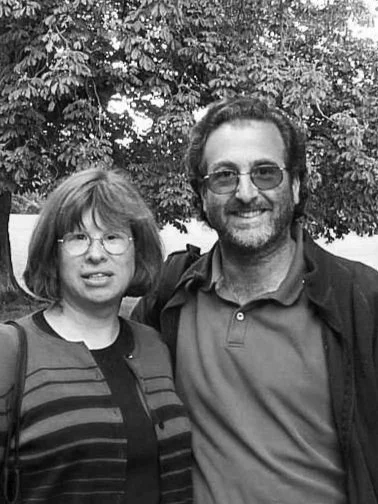 A black and white photo of a woman and man standing outdoors in front of large trees, smiling at the camera.