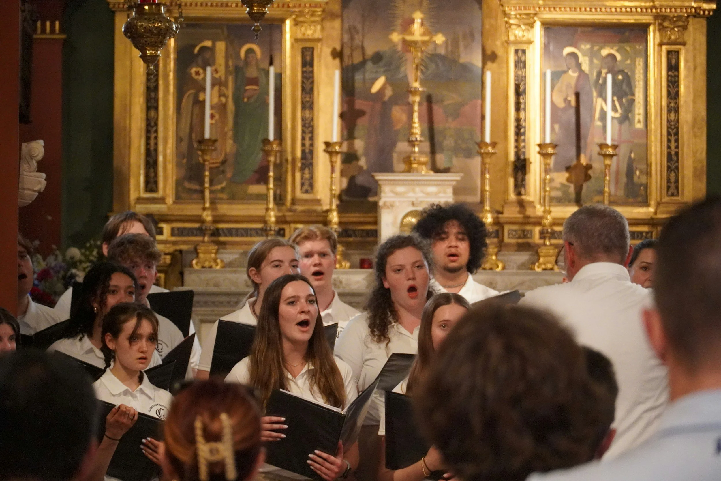 A choir of young people singing during a performance inside a church, with a gold ornate altar and religious paintings in the background.
