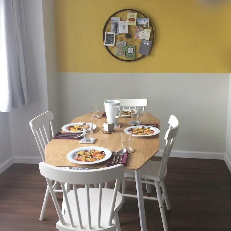 Wooden dining table set for a meal with white chairs, a yellow accent wall, and framed artwork in a bright, welcoming dining room.