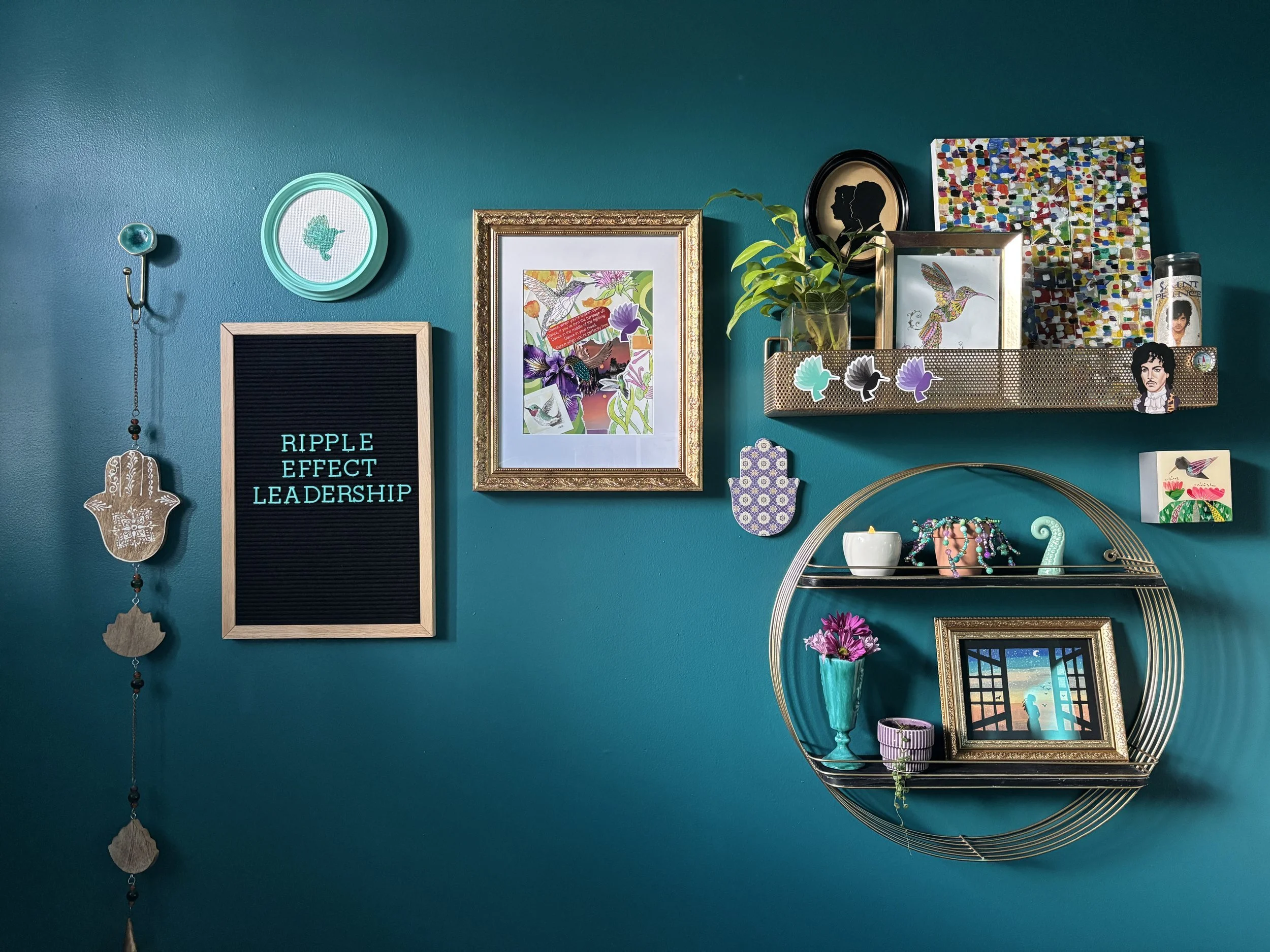 Dark teal wall with a gallery wall that includes gold accents, hummingbirds and a black letter board with turquoise letters that say Ripple Effect Leadership
