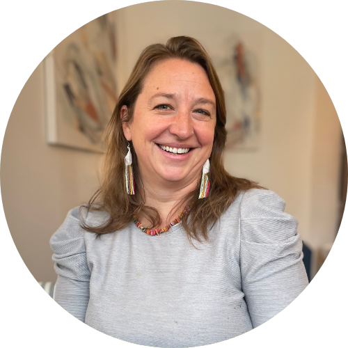 A woman with long brown hair, brown eyes, smiling in an indoor setting, with a blurred background of a room with a lamp and furniture.