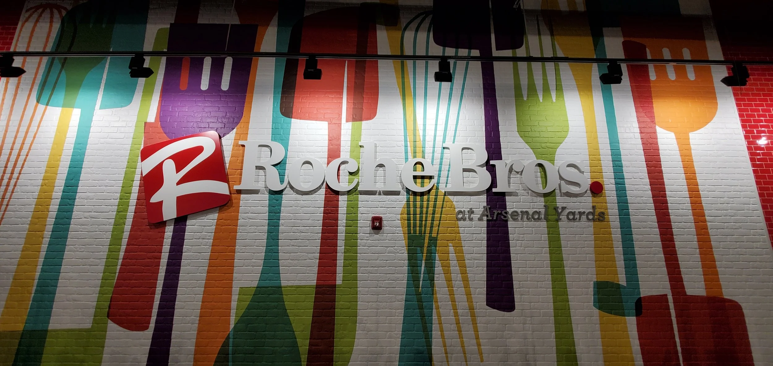 Decorative wall with colorful silhouettes of kitchen utensils such as forks, spoons, and whisks, and a black sign with the Roche Bros. logo and text 'at Arsenal Yards'.