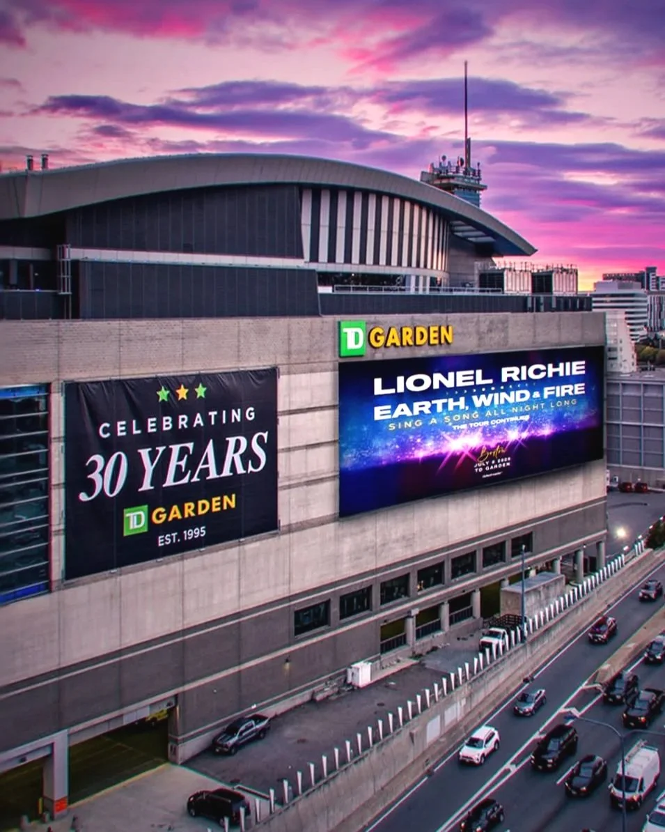Large urban building with billboard advertising celebrating 30 years of TD Garden, featuring a performance by Lionel Richie titled 'Earth, Wind & Fire.' City street with cars in the foreground at sunset with colorful sky.