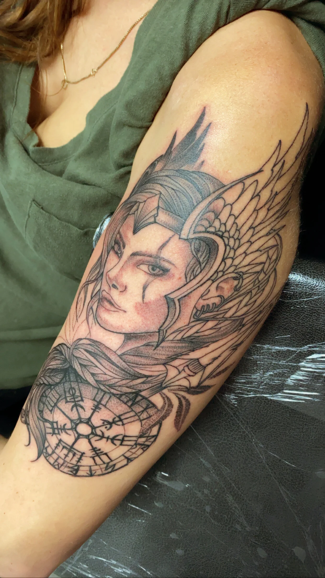 A tattoo on a person's upper arm depicting a woman's face with long flowing hair, wings, and a clock with Roman numerals.
