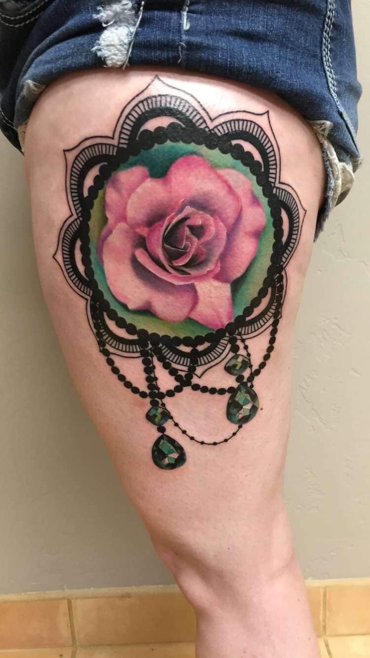 Tattoo of a pink and purple rose with green leaves surrounded by a black ornamental design resembling a lace pattern, with black beaded chains and green gem-like pendants hanging below.