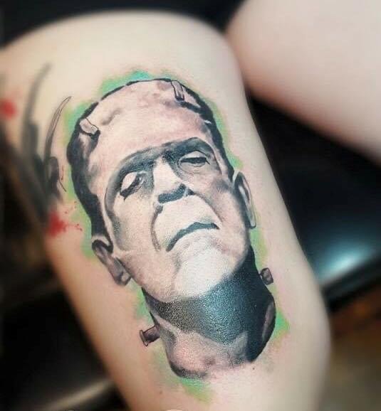 Tattoo of Frankenstein's monster on a person's arm, depicted in a realistic black and gray style with green and red accents around the head.